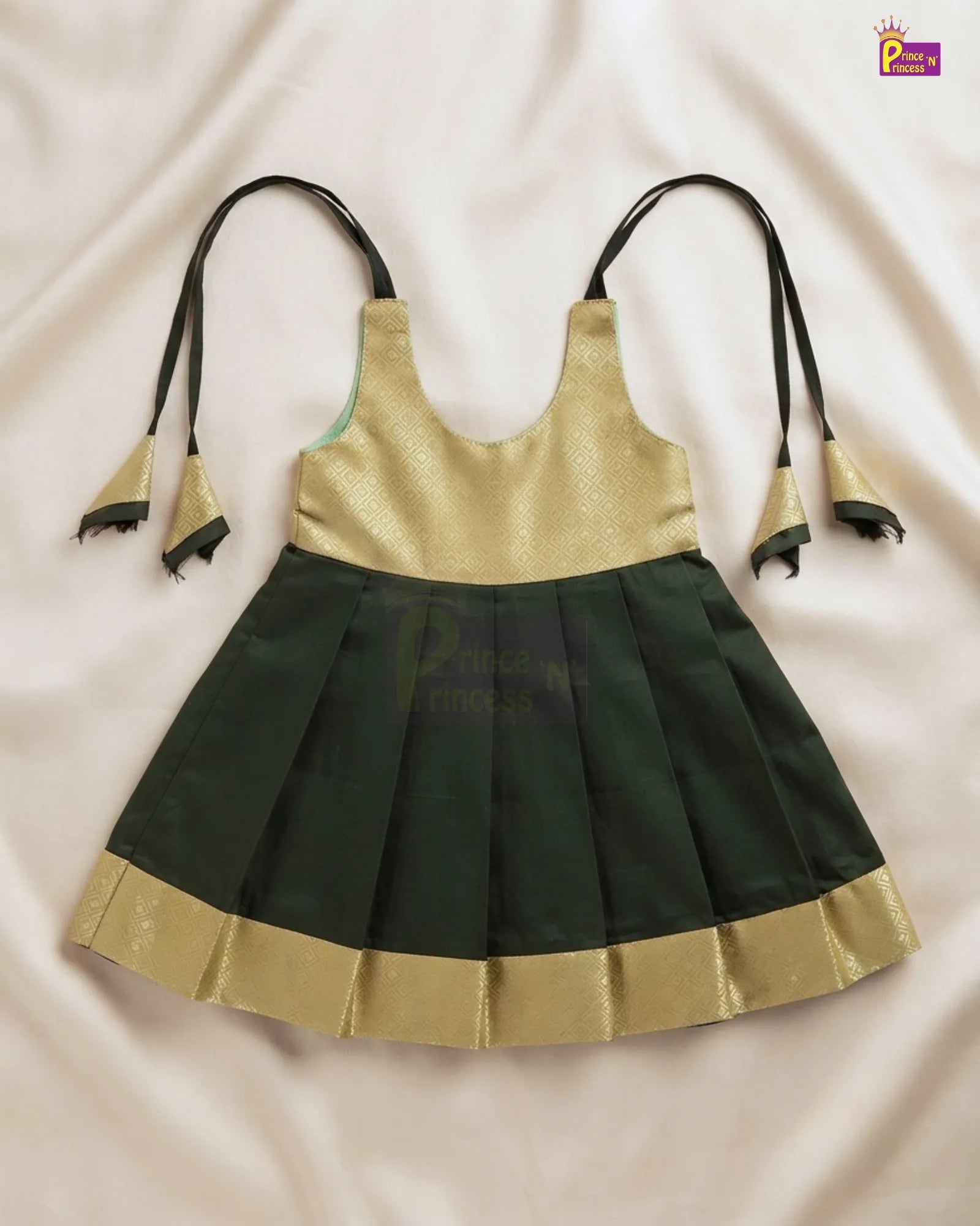 A children's sleeveless dress with a metallic gold upper bodice and a dark green pleated skirt, laid flat on a light beige fabric background. The dress has thin black shoulder ties with gold and black tassels and a gold border along the skirt hem.