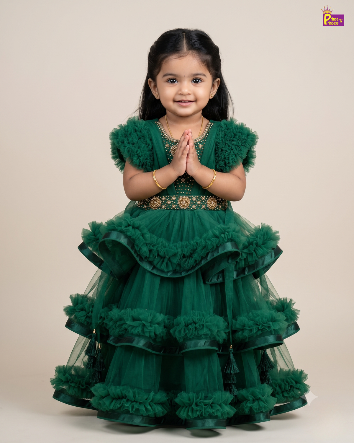 A young girl with dark hair is wearing a green tiered tulle party dress with ruffled trim and gold embroidery, standing against a plain beige background with her hands together in a prayer pose.