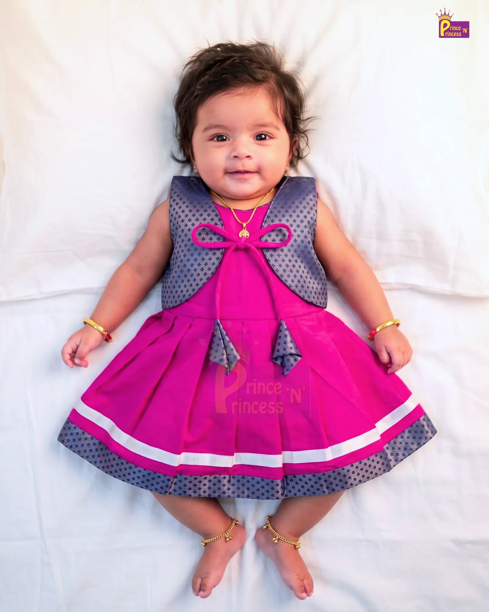 A baby girl lying on a white sheet wearing a bright pink and gray polka dot dress with gold jewelry.