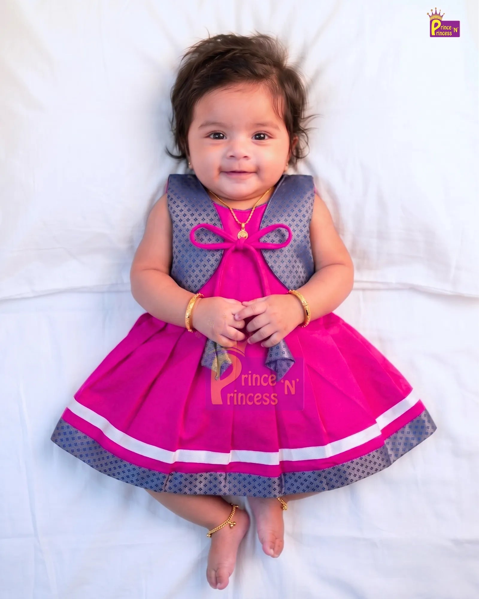 A baby girl lying on a white sheet wearing a bright pink and gray printed dress with gold jewelry. The dress features a gray patterned bodice with a pink bow at the neckline, a matching pink skirt with white trim, and the text 'Princess N' printed on the front.