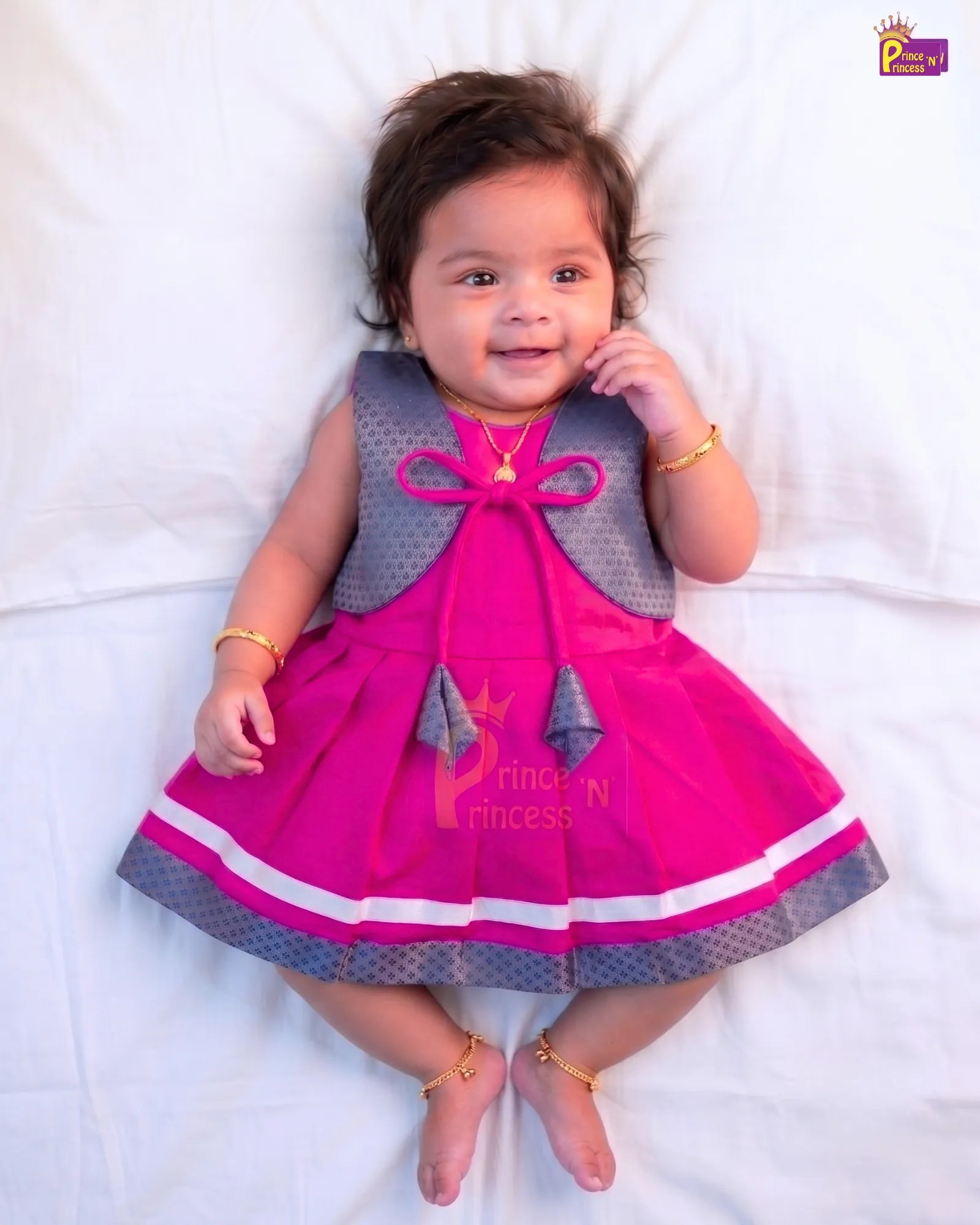 A baby girl lying on a white sheet, wearing a bright pink and gray dress with gold jewelry.