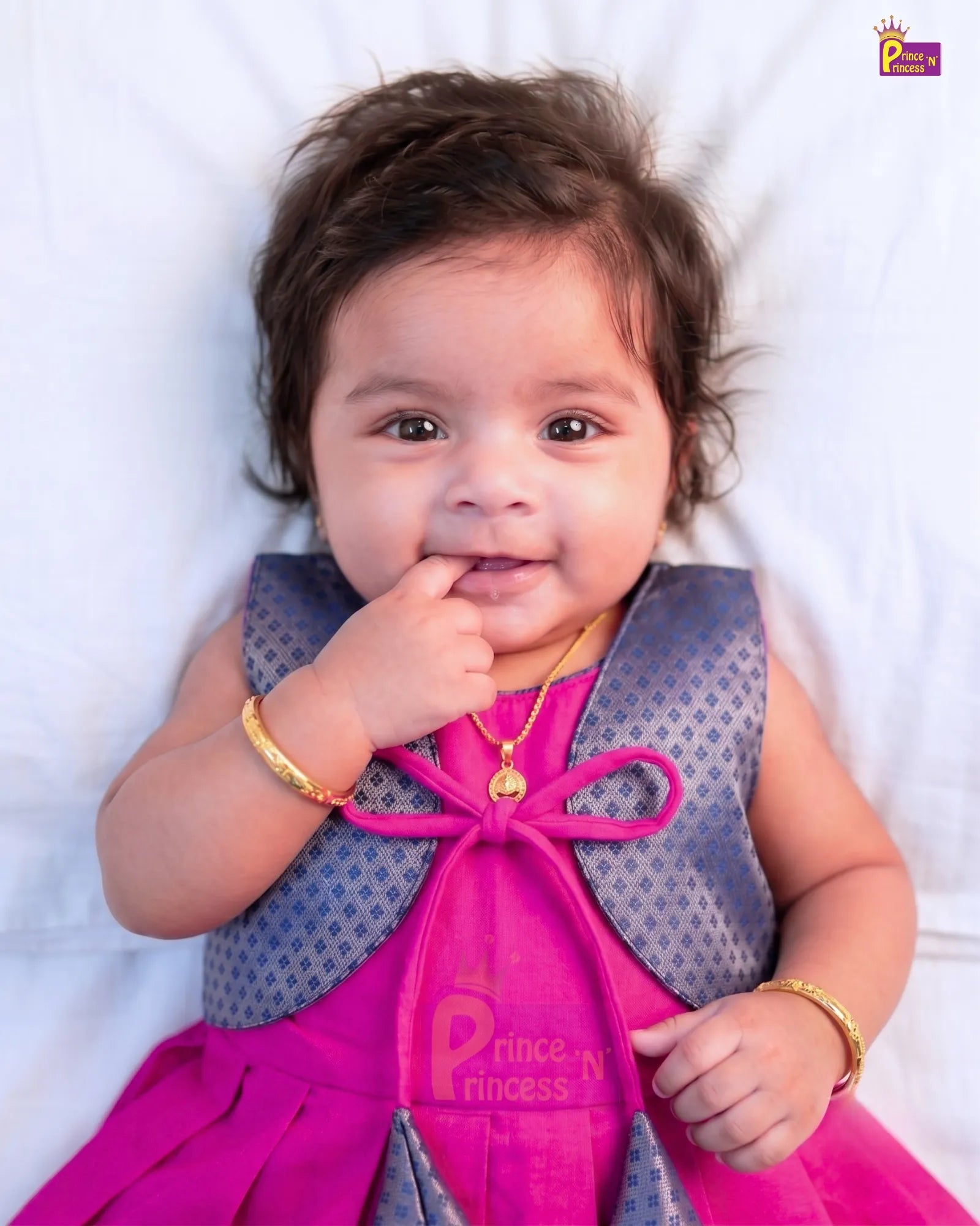 A baby lying on a white blanket wearing a bright pink and purple traditional outfit with gold jewelry. The brand logo 'Prince N Princess' is visible in the top right corner.