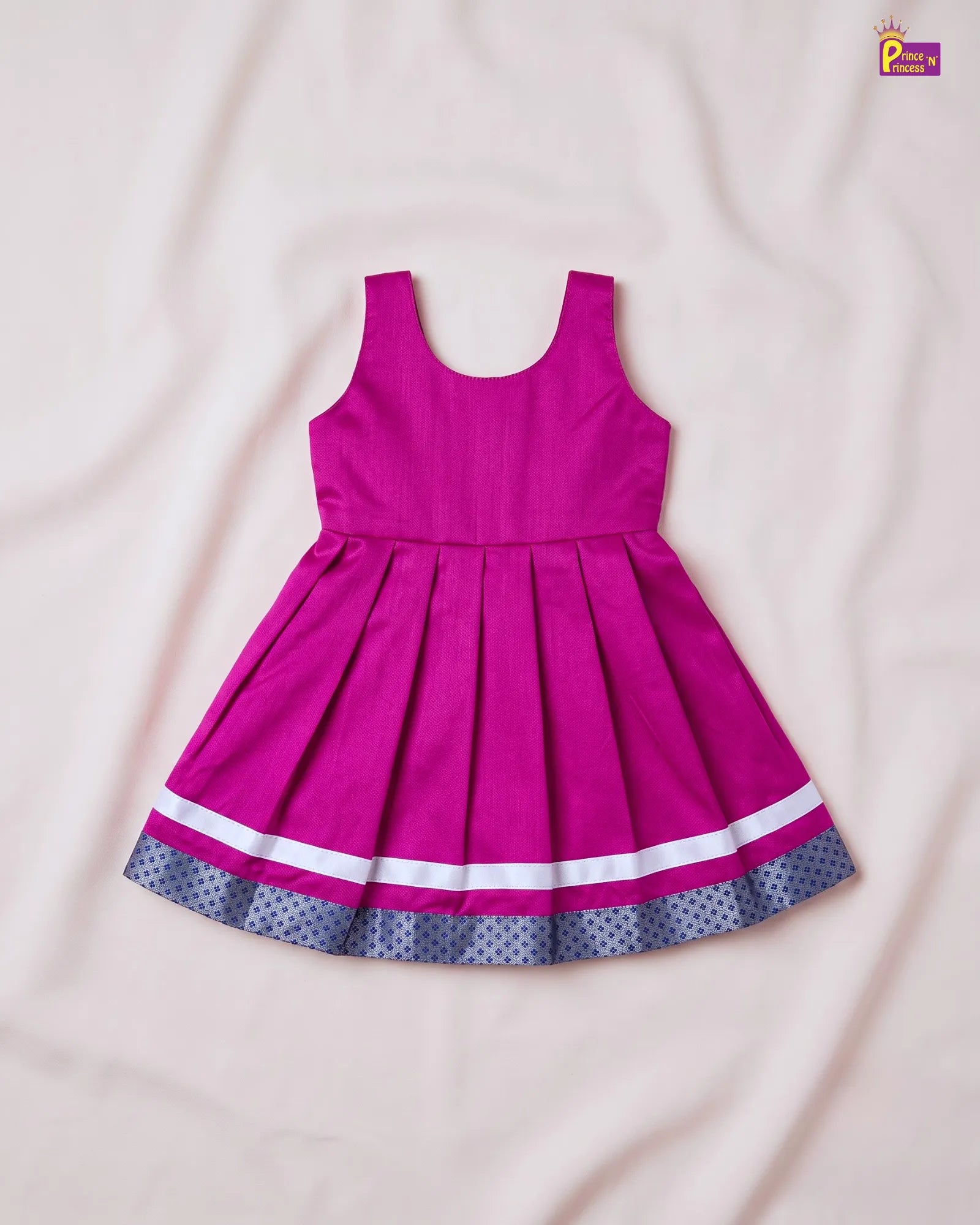 A sleeveless magenta pink children's dress with a pleated skirt, featuring a white stripe and a navy blue polka dot border on the hem, laid flat on a light pink fabric background.