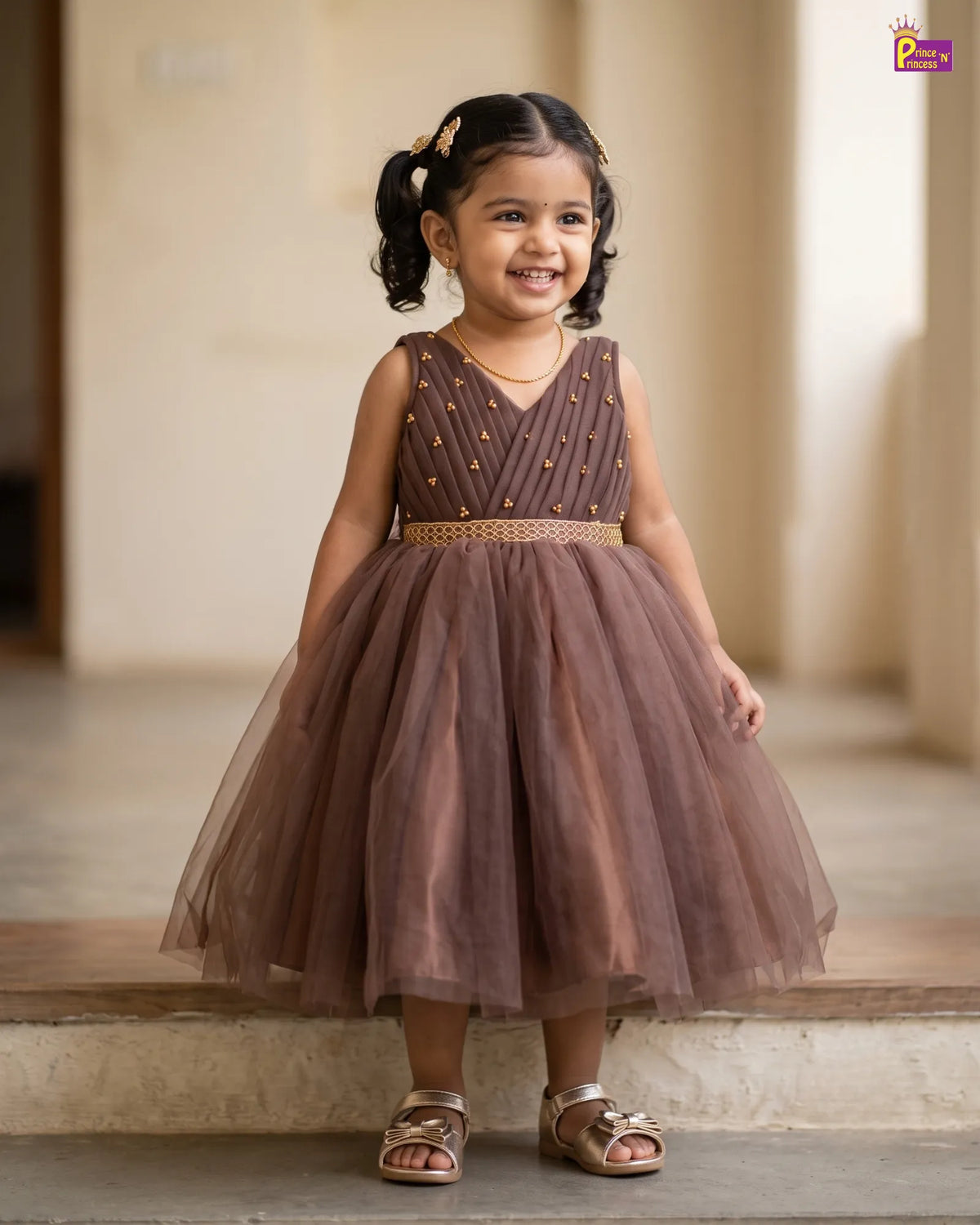 A young girl with dark hair in pigtails stands smiling on stone steps, wearing a sleeveless brown tulle dress with gold embellishments and sandals.