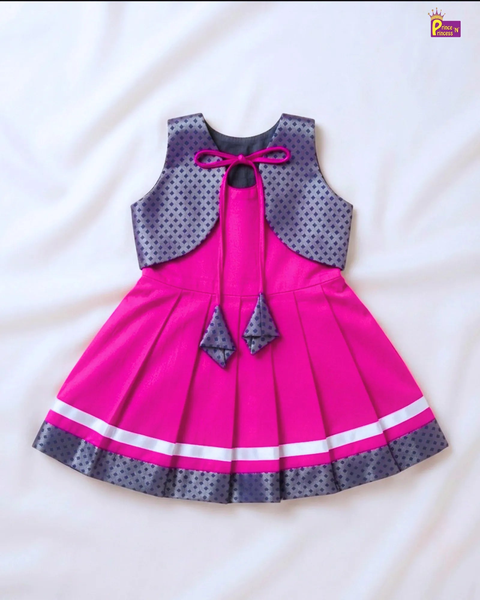 A sleeveless children's dress laid flat on a white fabric background. The dress has a bright pink pleated skirt and a dark gray polka-dot sleeveless overlay with a pink bow at the neckline.