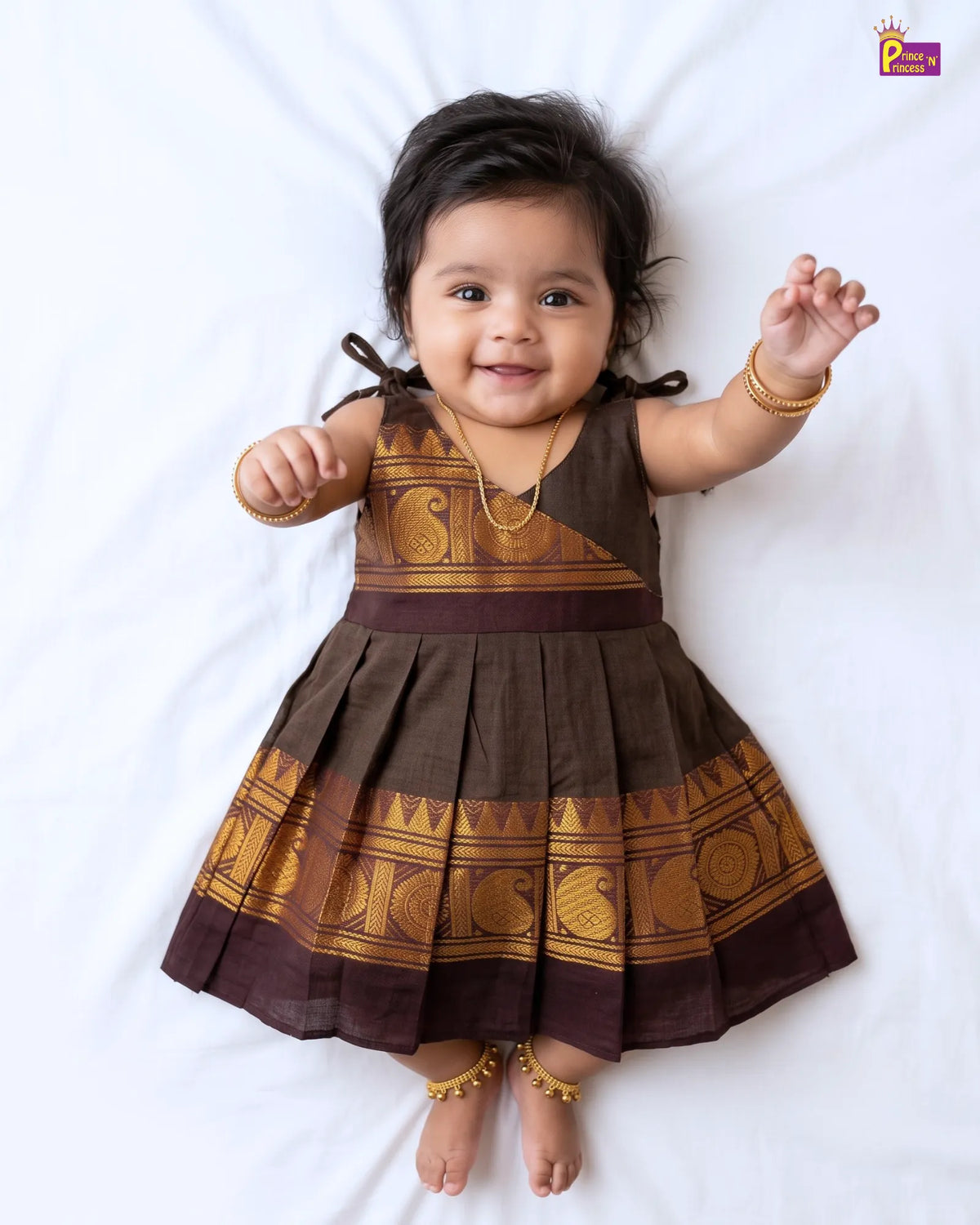 A baby girl lying on a white sheet wearing a brown and gold printed traditional Indian dress with gold ankle bracelets and a gold necklace.