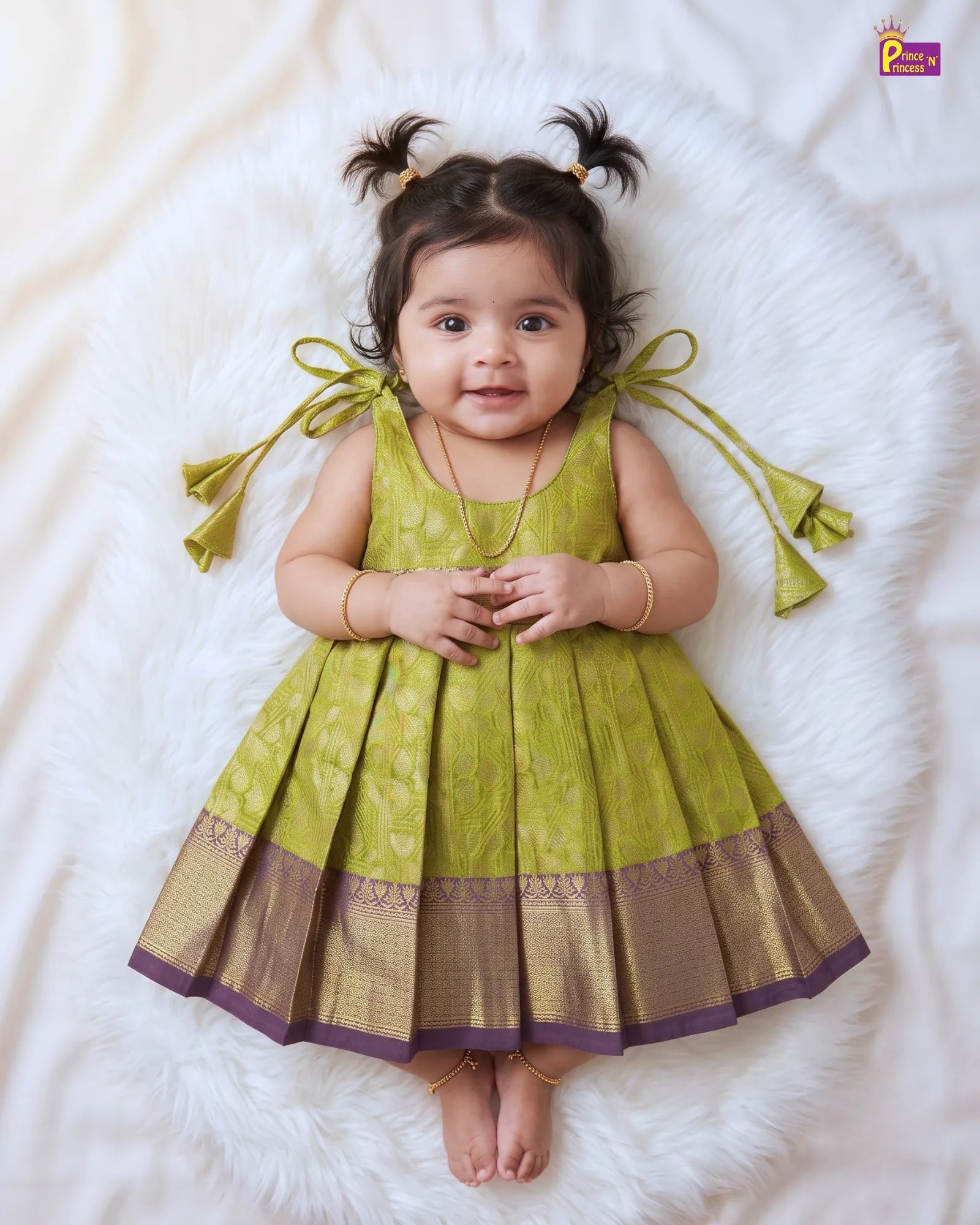A toddler girl lying on a white faux fur rug wearing a sleeveless olive green and gold pleated dress with purple border trim and matching green ribbon bows on the shoulders.