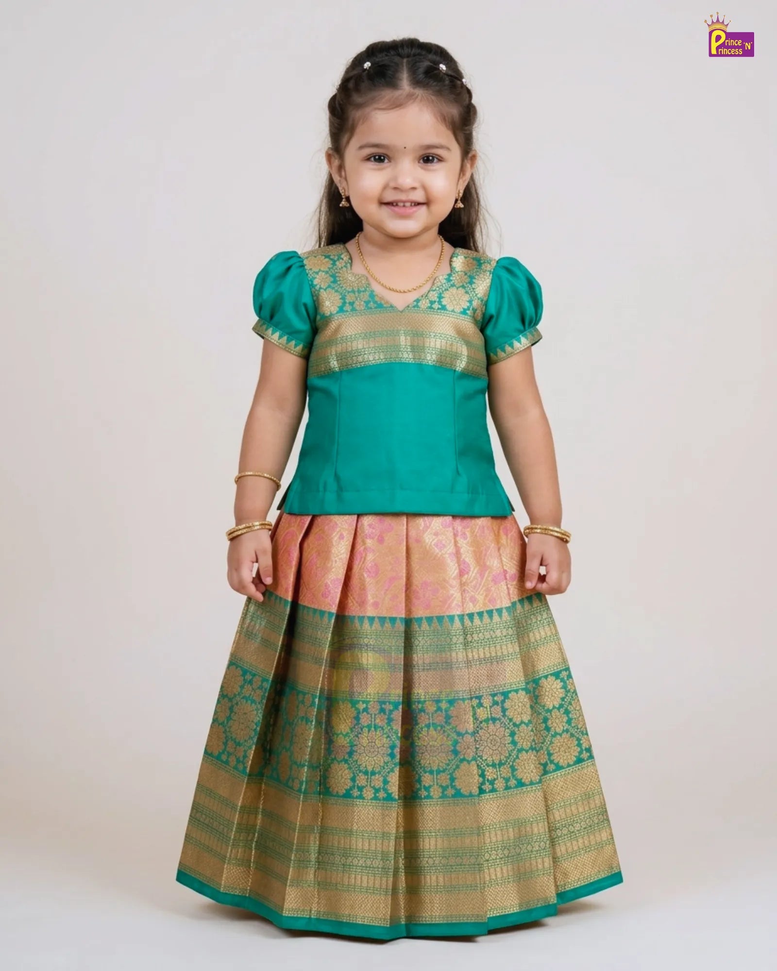 A young girl modeling a traditional Indian ethnic outfit consisting of a teal green top with gold zari embroidery and a matching pleated skirt in pink and green with gold patterns, set against a plain light gray background.