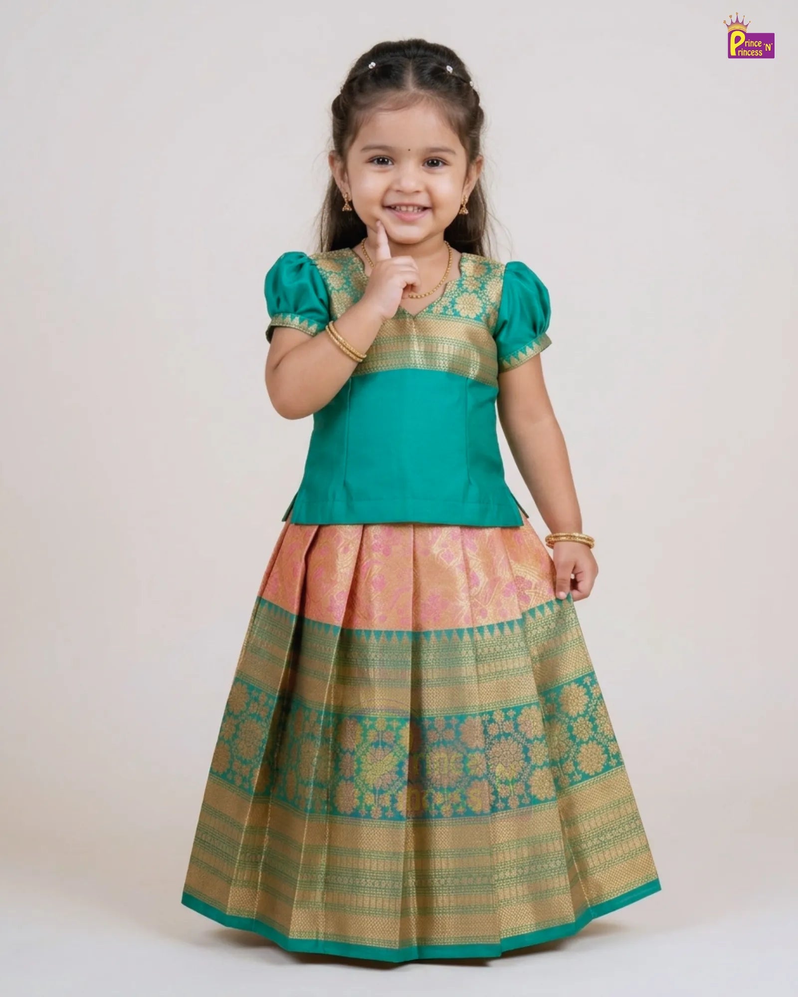 A young girl modeling a traditional Indian ethnic dress with a teal green top and a pleated skirt featuring gold and pink zari patterns, set against a plain light beige background.