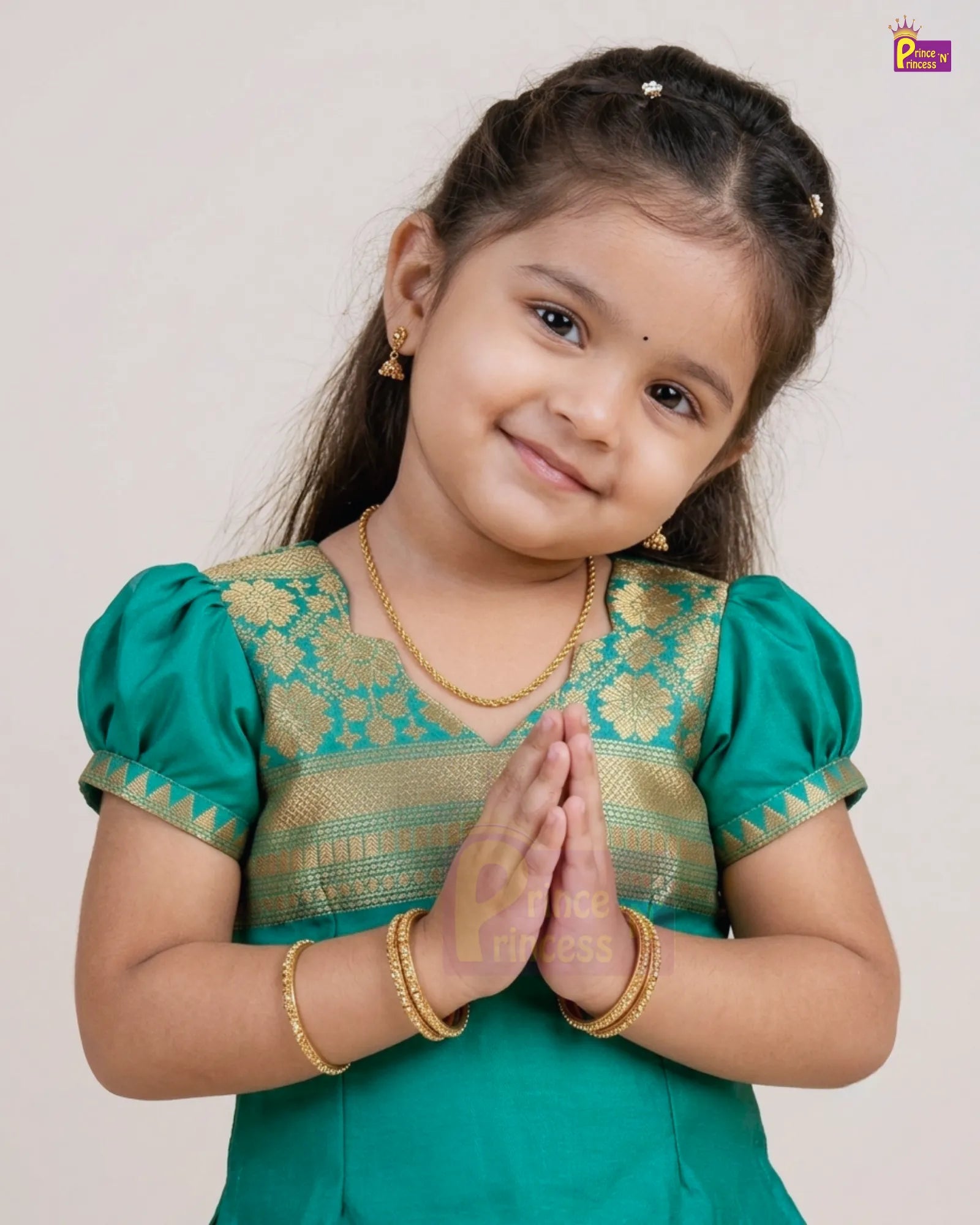 A young girl with dark hair styled in braids, wearing a traditional green and gold embroidered Indian outfit with gold jewelry, posing with her hands together against a plain light background.