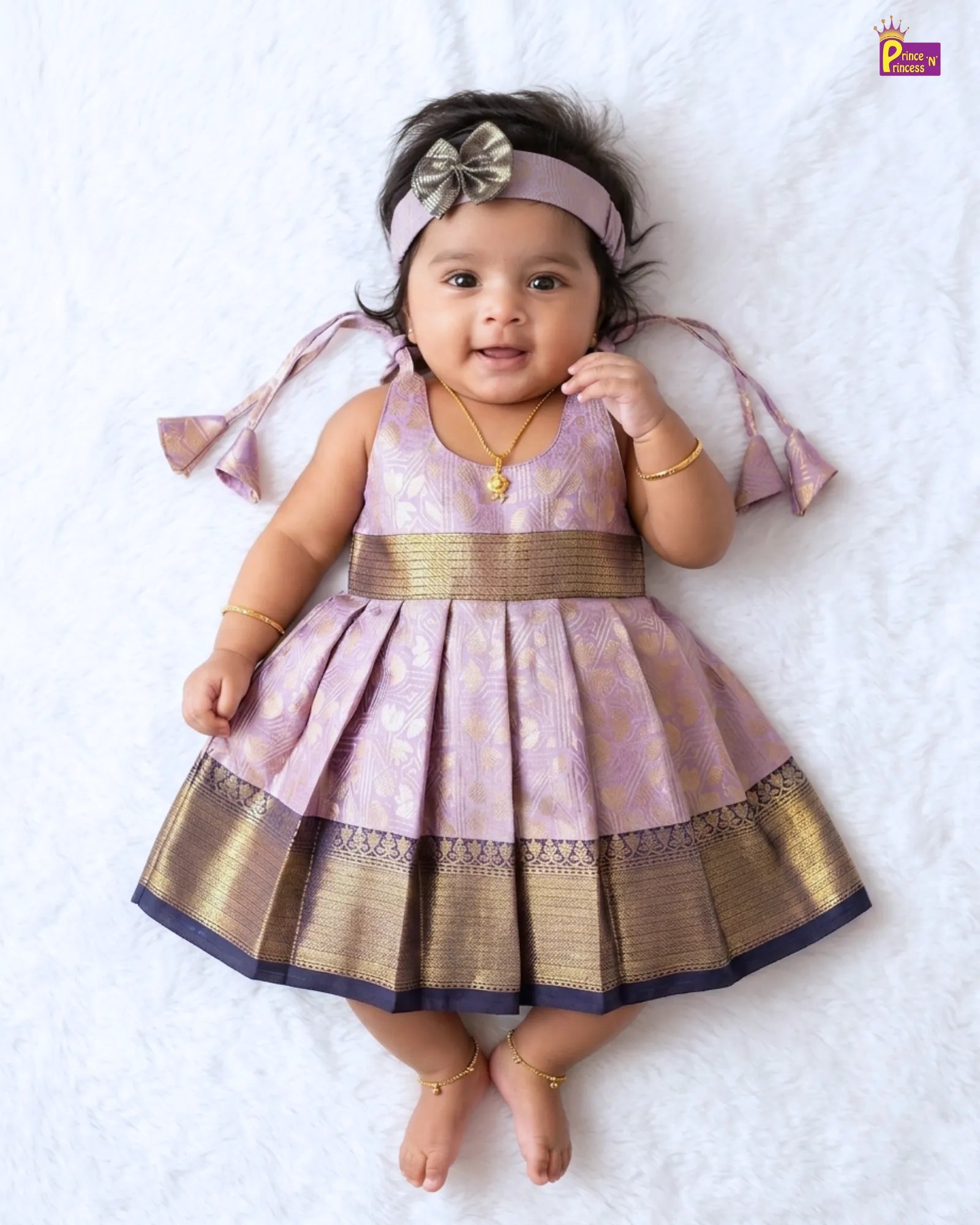 A baby girl wearing a traditional Indian lavender and gold Banarasi dress with a matching headband, lying on a white textured blanket.