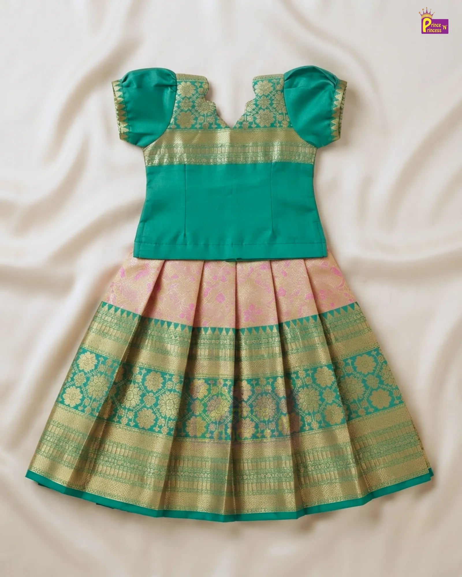 A traditional Indian girl's dress with a solid teal green top and a pleated skirt featuring pink, gold, and teal green patterns, laid flat on a cream-colored fabric background.