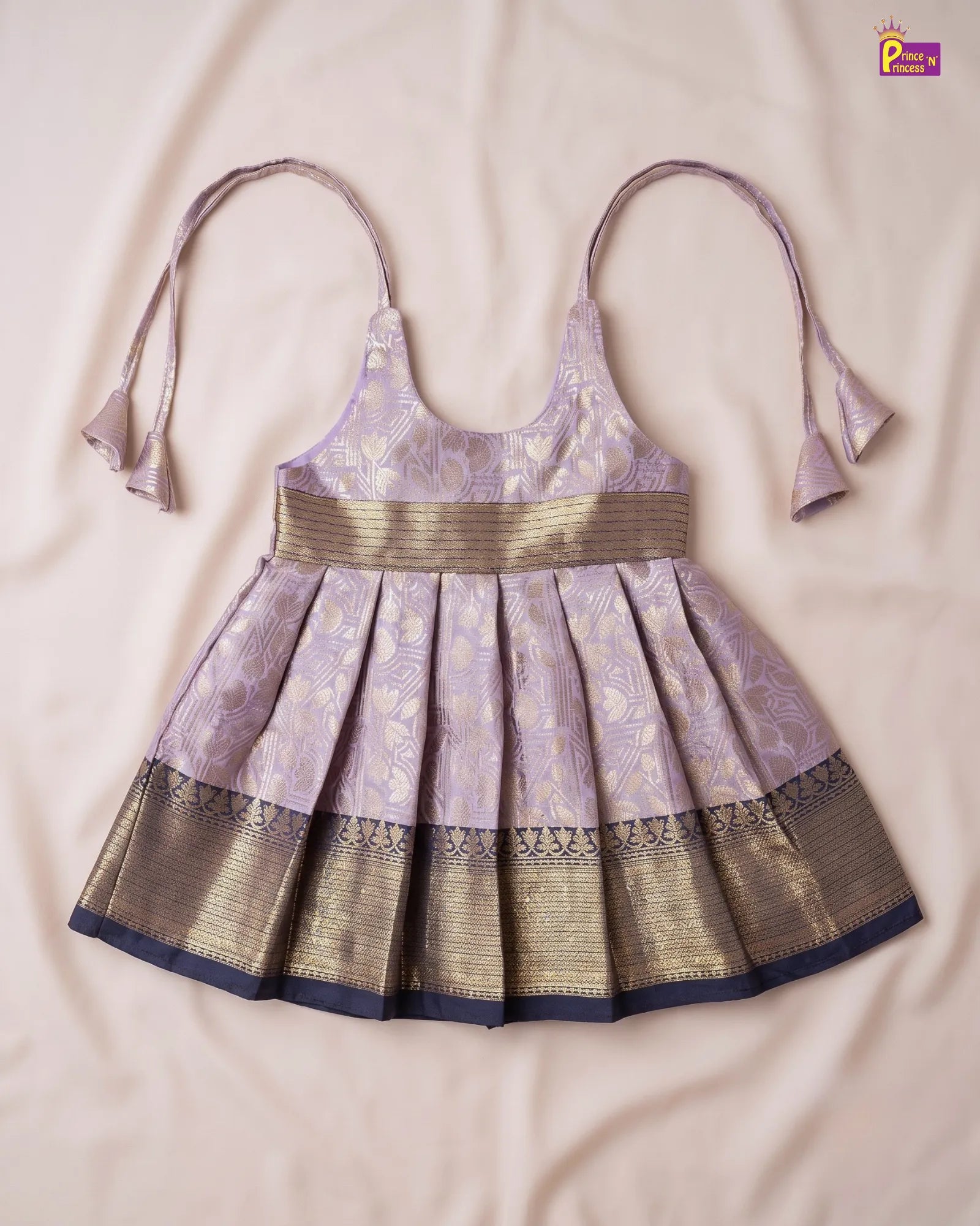 A traditional Indian toddler girl's salwar kameez dress in lavender and gold, laid flat on a beige fabric background. The dress features a lavender body with gold floral patterns, a wide gold waistband, and a pleated skirt with a gold zari border and navy blue trim.