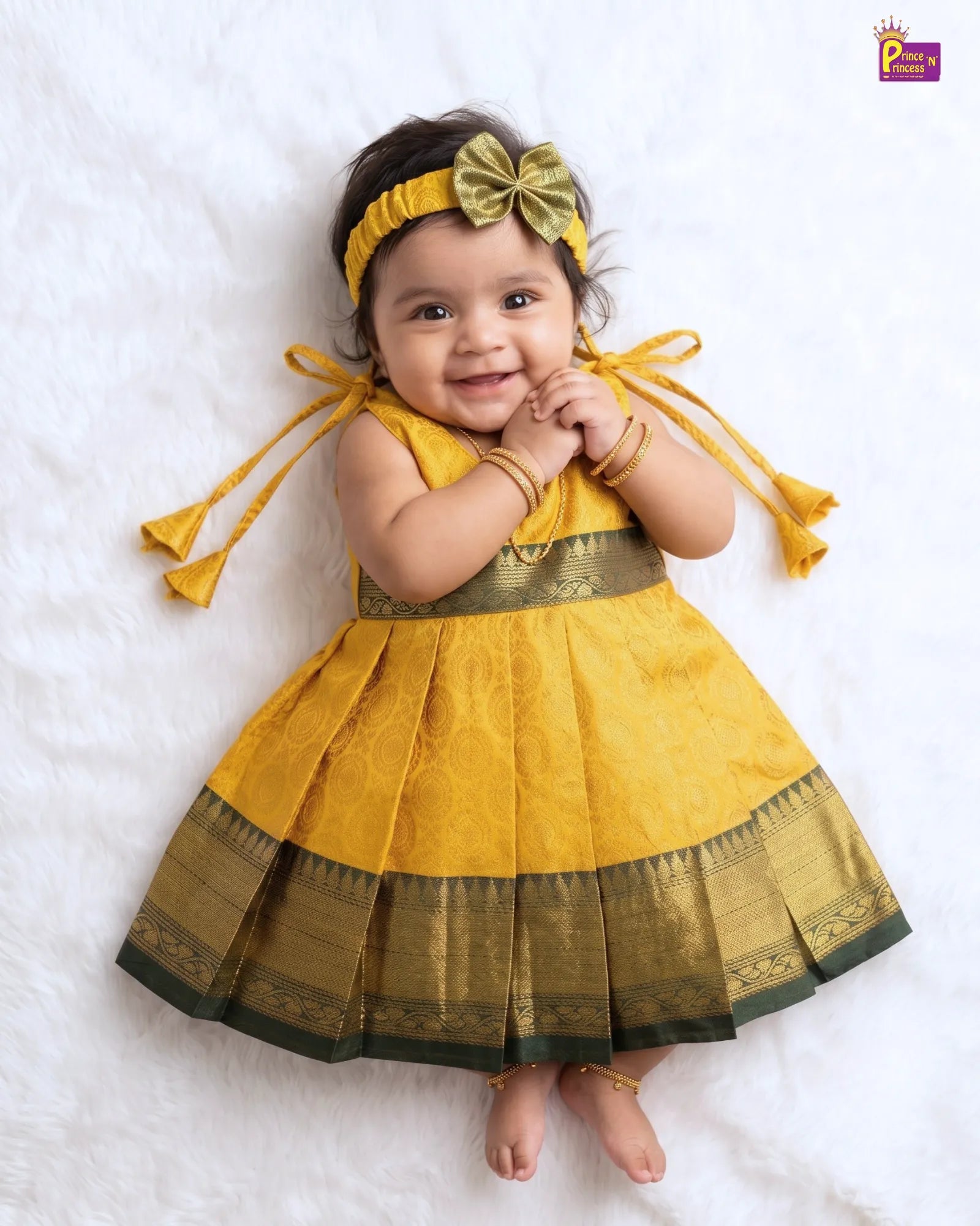 A baby girl lying on a white textured blanket, wearing a yellow and gold traditional dress with a matching bow headband and gold bangles.