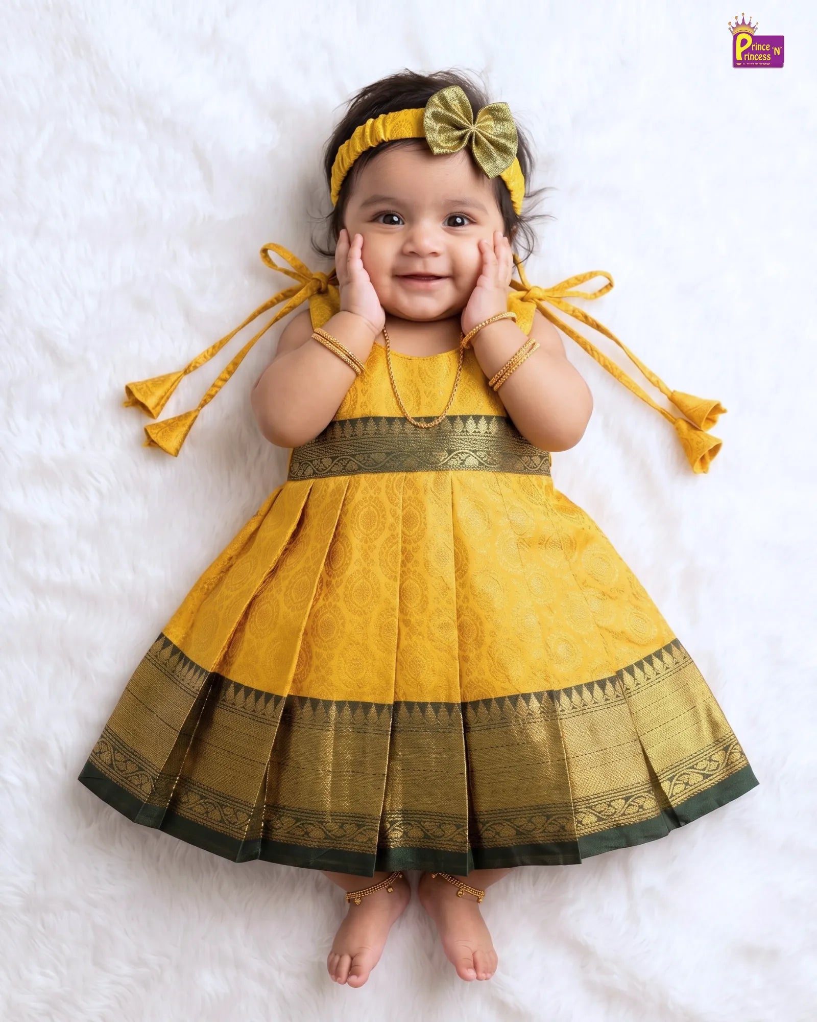 A baby girl lying on a white faux fur surface, wearing a yellow and gold traditional Indian dress with a pleated skirt, a matching yellow headband with a green bow, and gold jewelry.