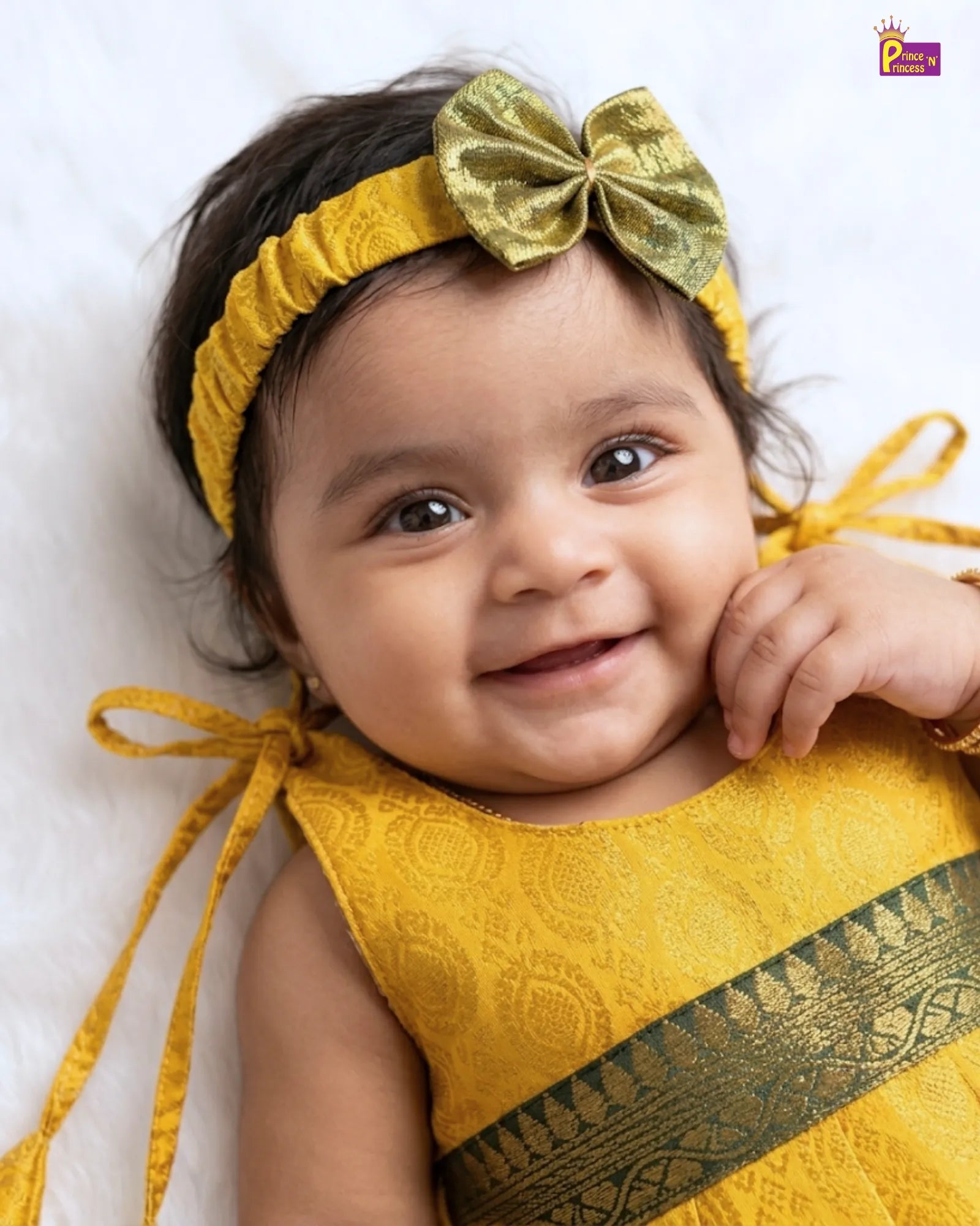 A smiling baby wearing a yellow textured dress with a dark green patterned sash and a matching yellow headband with a metallic gold bow, lying on a white textured blanket.
