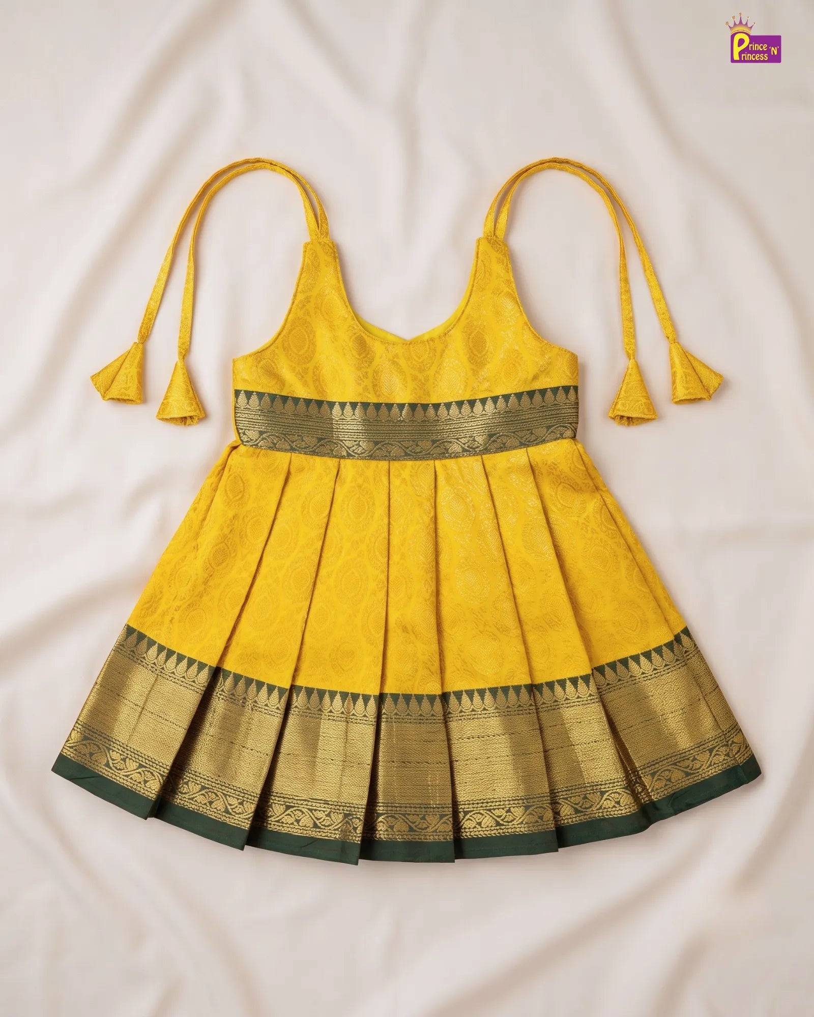 A yellow sleeveless baby dress with gold and green zari borders, laid flat on a white fabric background. The dress features thin shoulder ties with tassel ends and a pleated skirt.