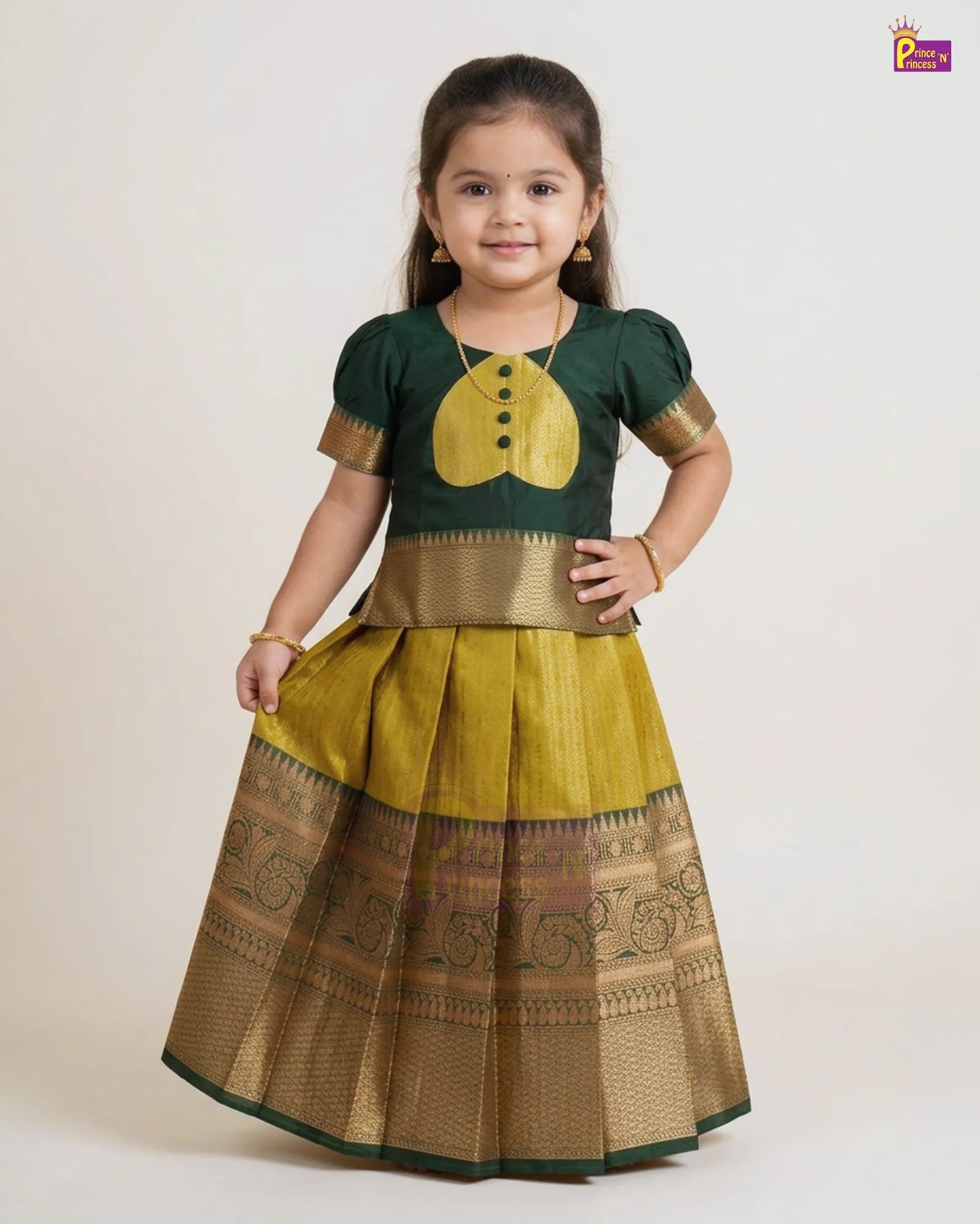 A young girl modeling a traditional Indian ethnic wear set consisting of a dark green short-sleeve blouse with a yellow chest motif and gold trim, paired with a pleated yellow skirt featuring a wide gold zari border.