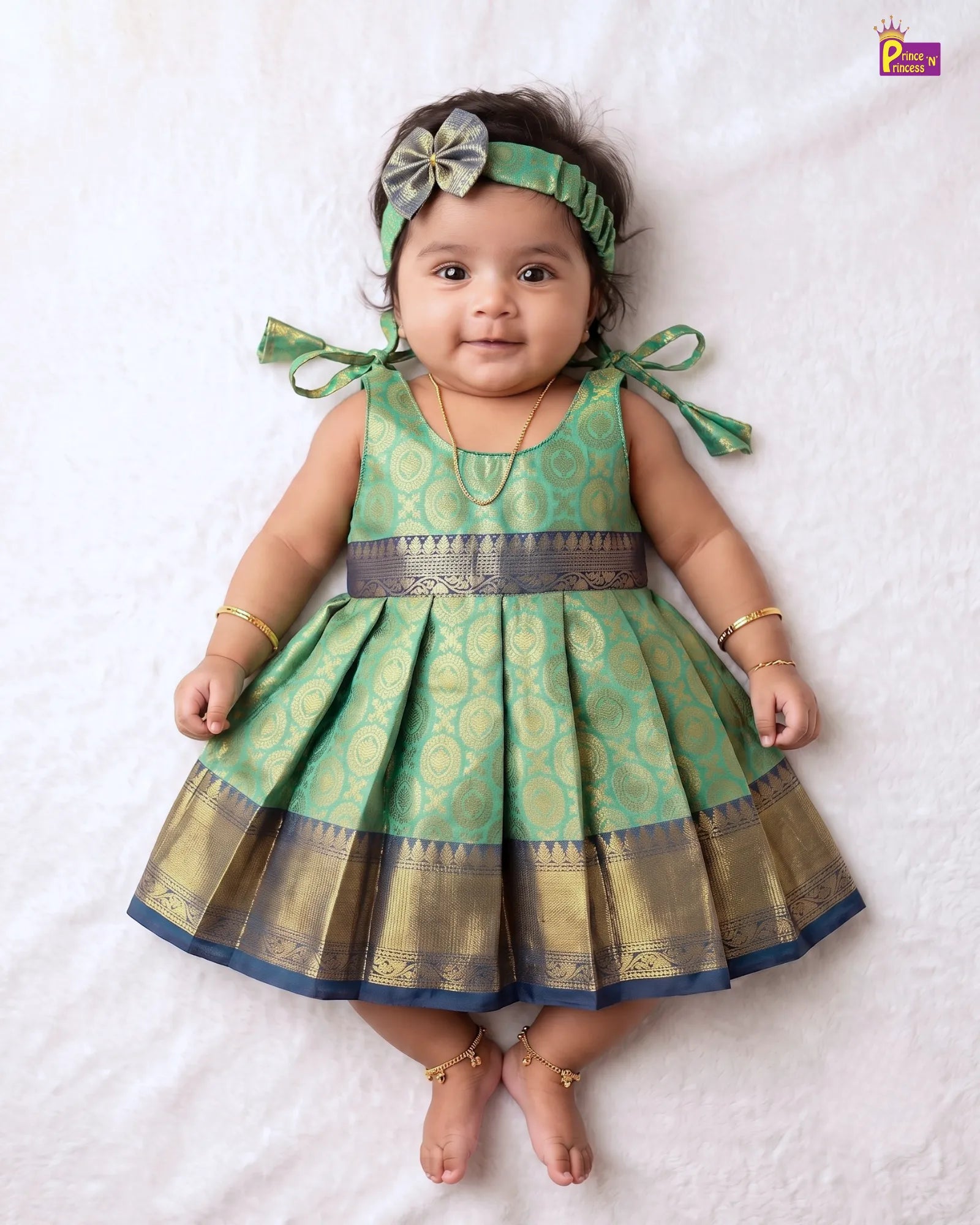 A baby girl wearing a green and gold patterned sleeveless dress with a matching headband, lying on a white textured blanket.