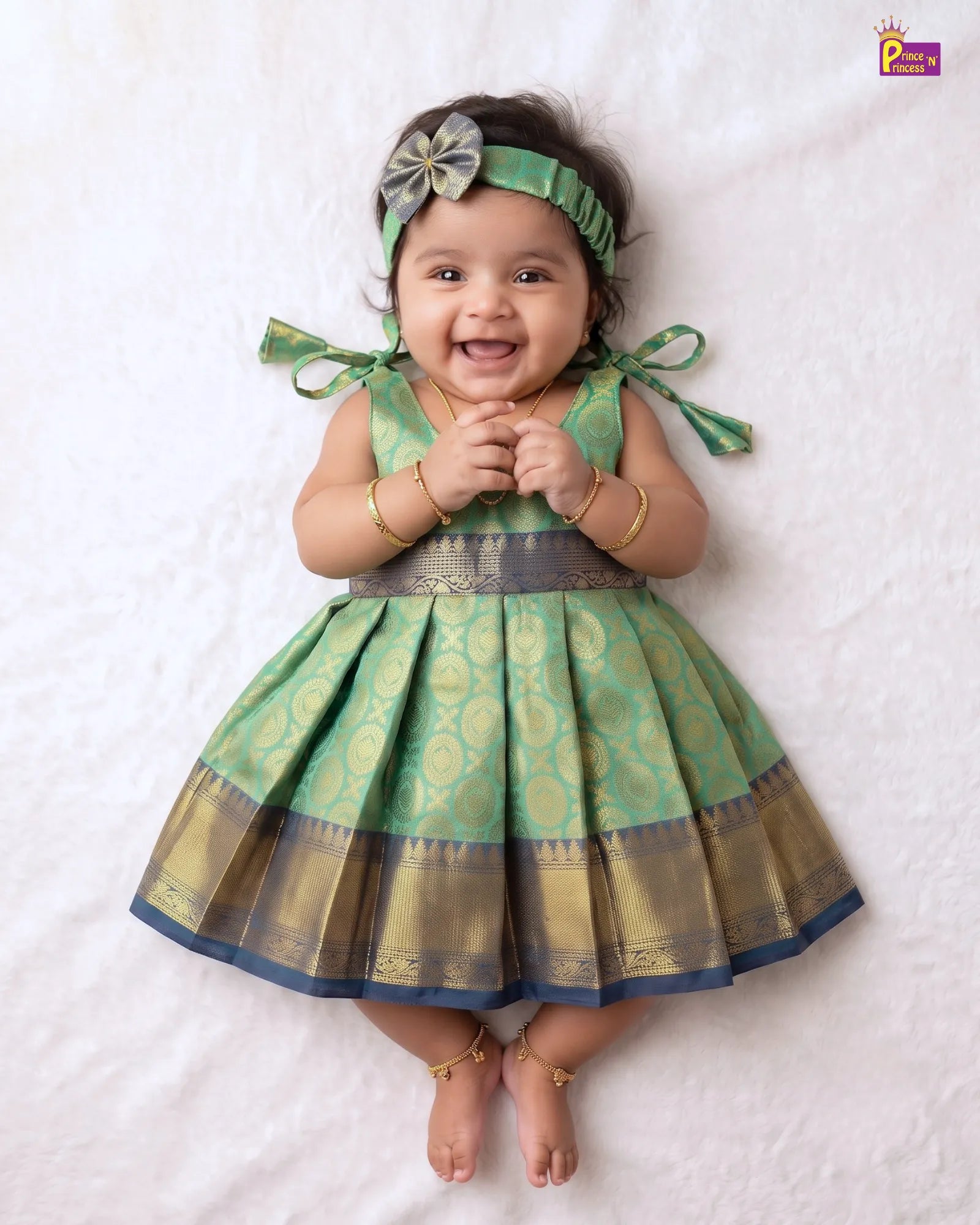 A baby girl lying on a white textured blanket, wearing a green and gold patterned traditional dress with a matching green headband and gold bangles.