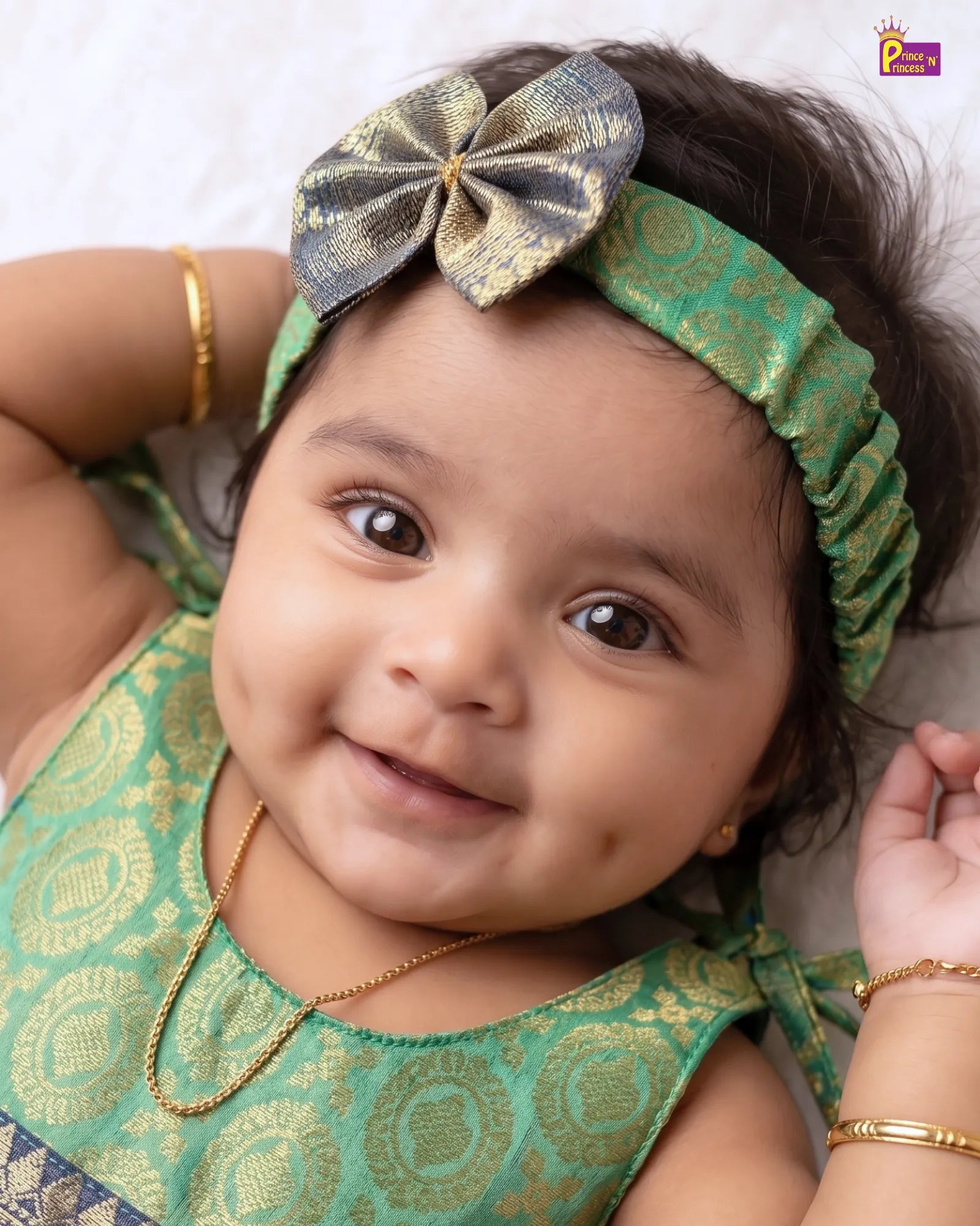 A close-up of a smiling baby wearing a green and gold patterned traditional outfit, a matching green and gold bow headband, gold jewelry, and lying on a light textured surface.