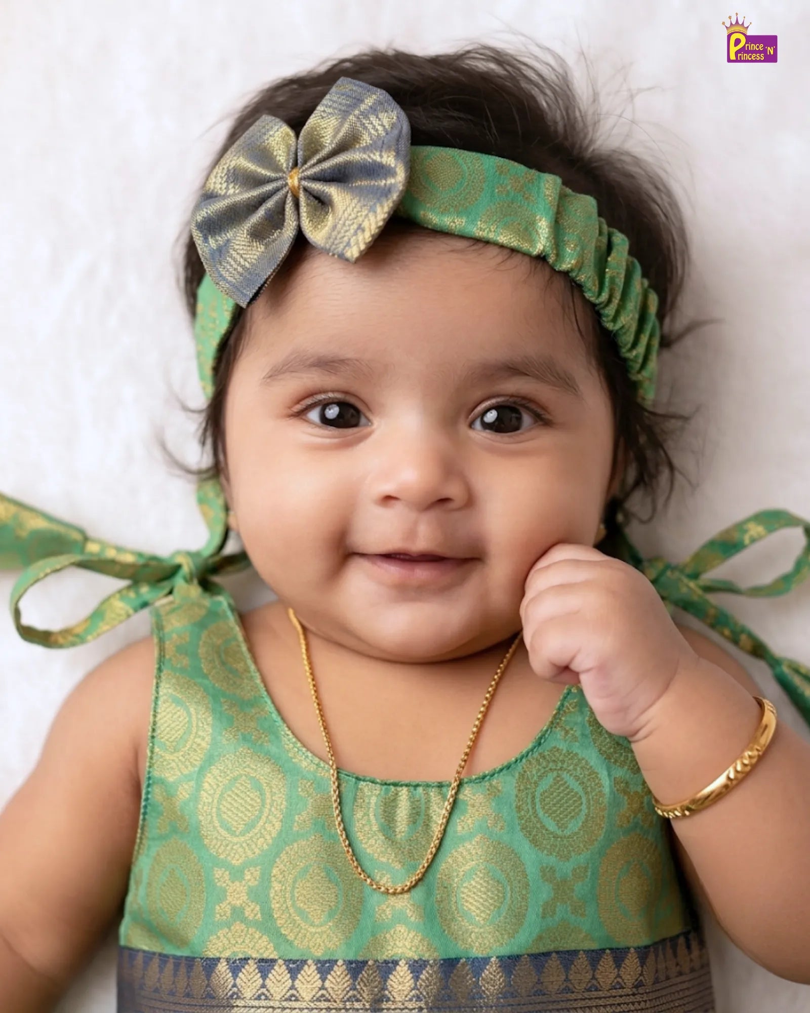 A baby girl wearing a green and gold patterned traditional top, a matching green headband with a large bow, a gold necklace, and a gold bracelet, lying down against a white textured background.