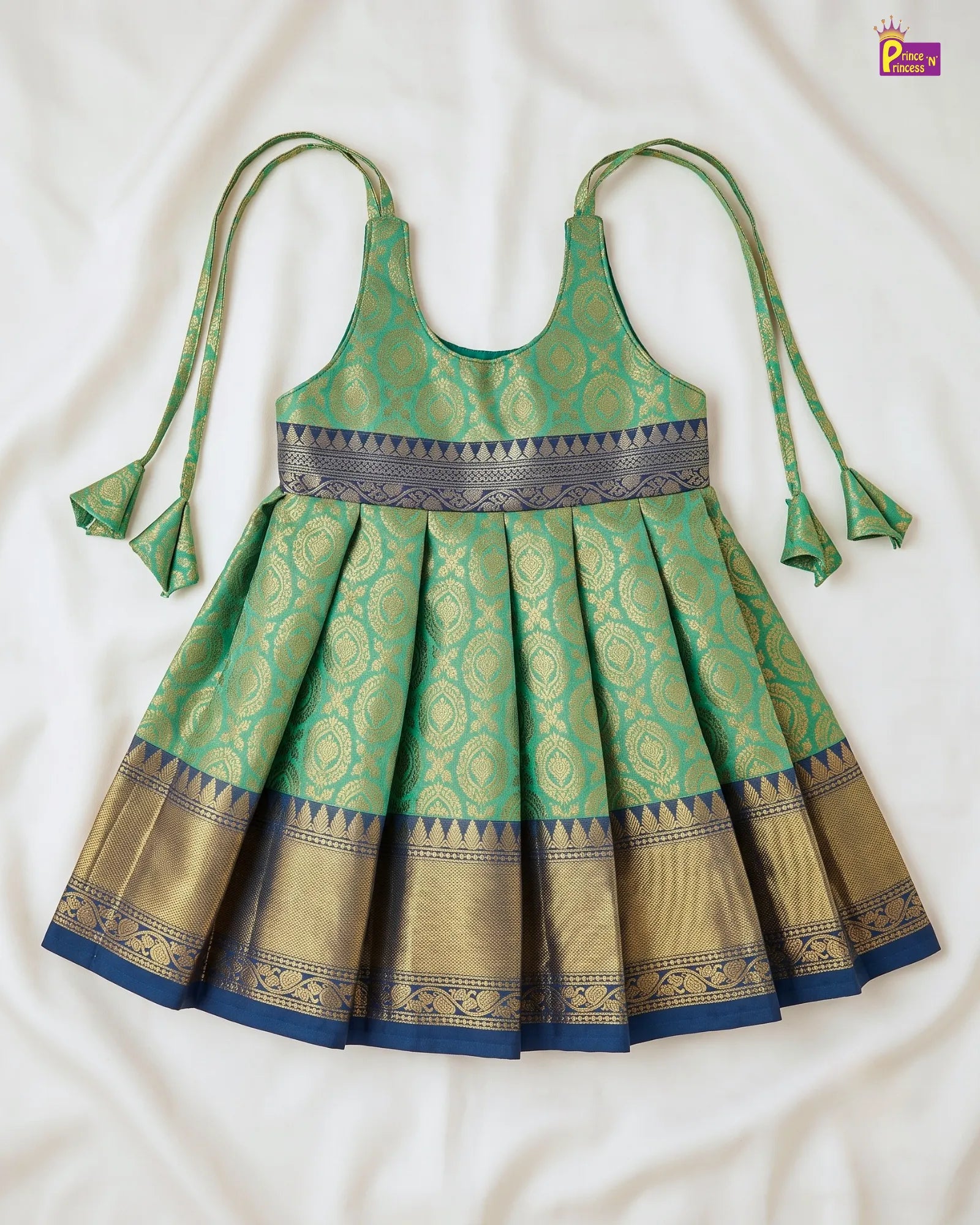 A green and gold Banarasi child's dress with tie-shoulder straps, laid flat on a white fabric background.