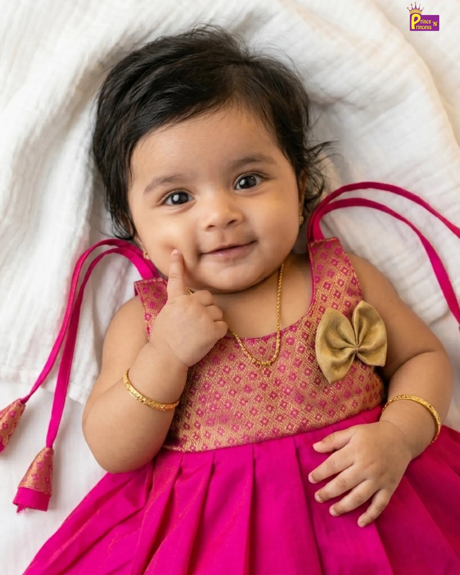 Baby in a pink dress with a gold bow, lying on a white blanket