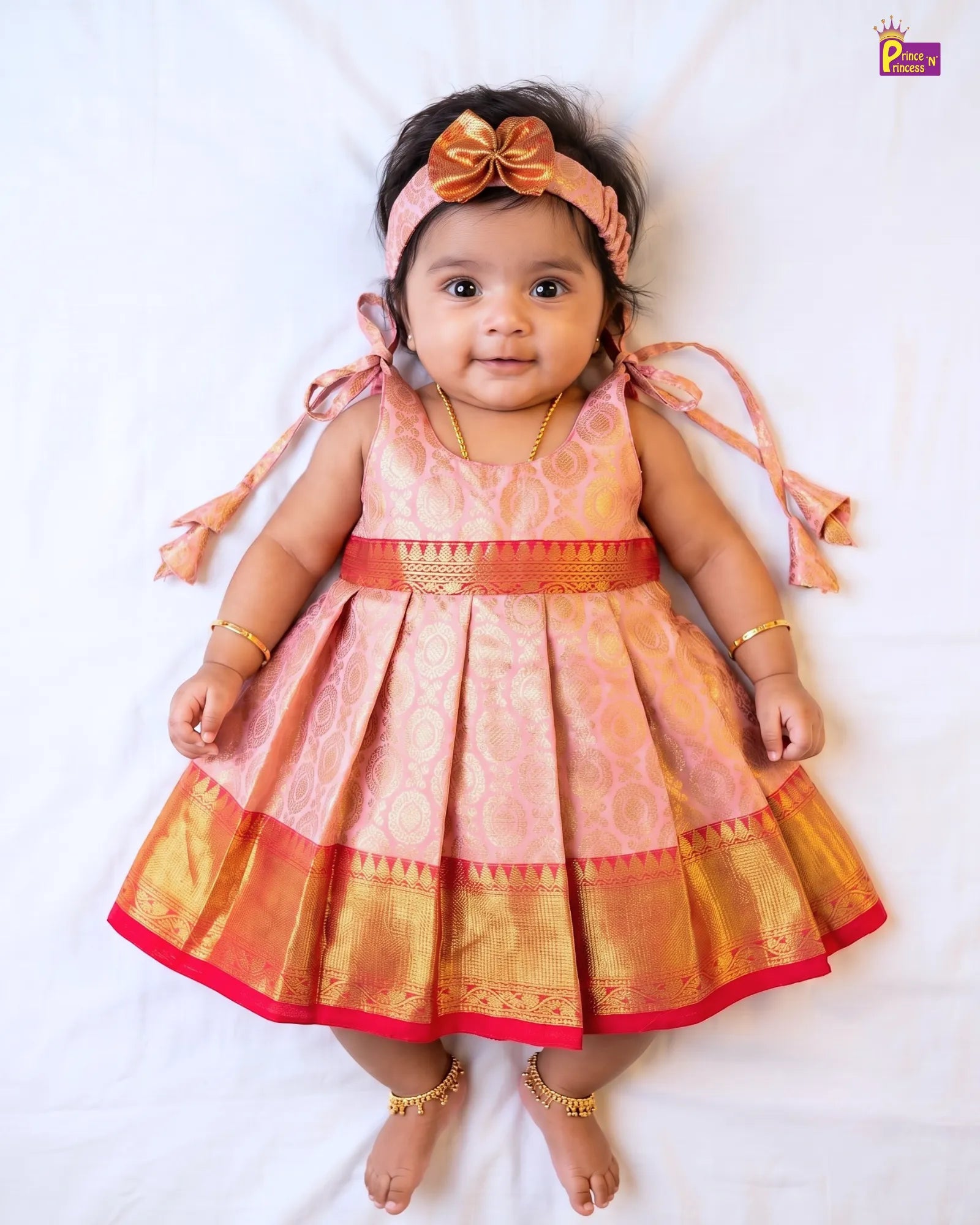 A baby girl wearing a traditional Indian salwar kameez dress in pink and gold with a matching pink bow headband, lying on a white sheet.