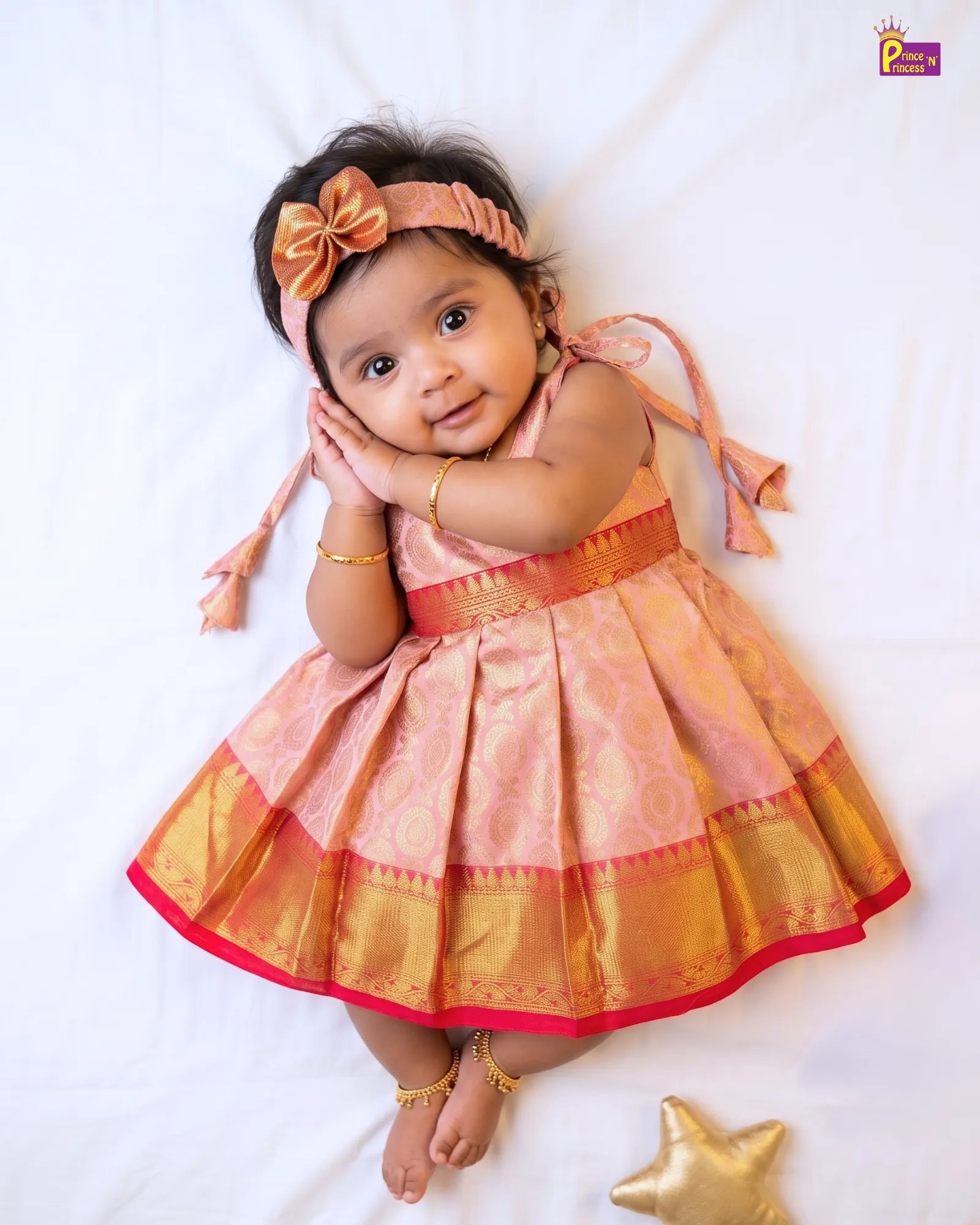 A baby girl lying on a white sheet wearing a peach and gold traditional Indian dress with a matching headband and gold jewelry.