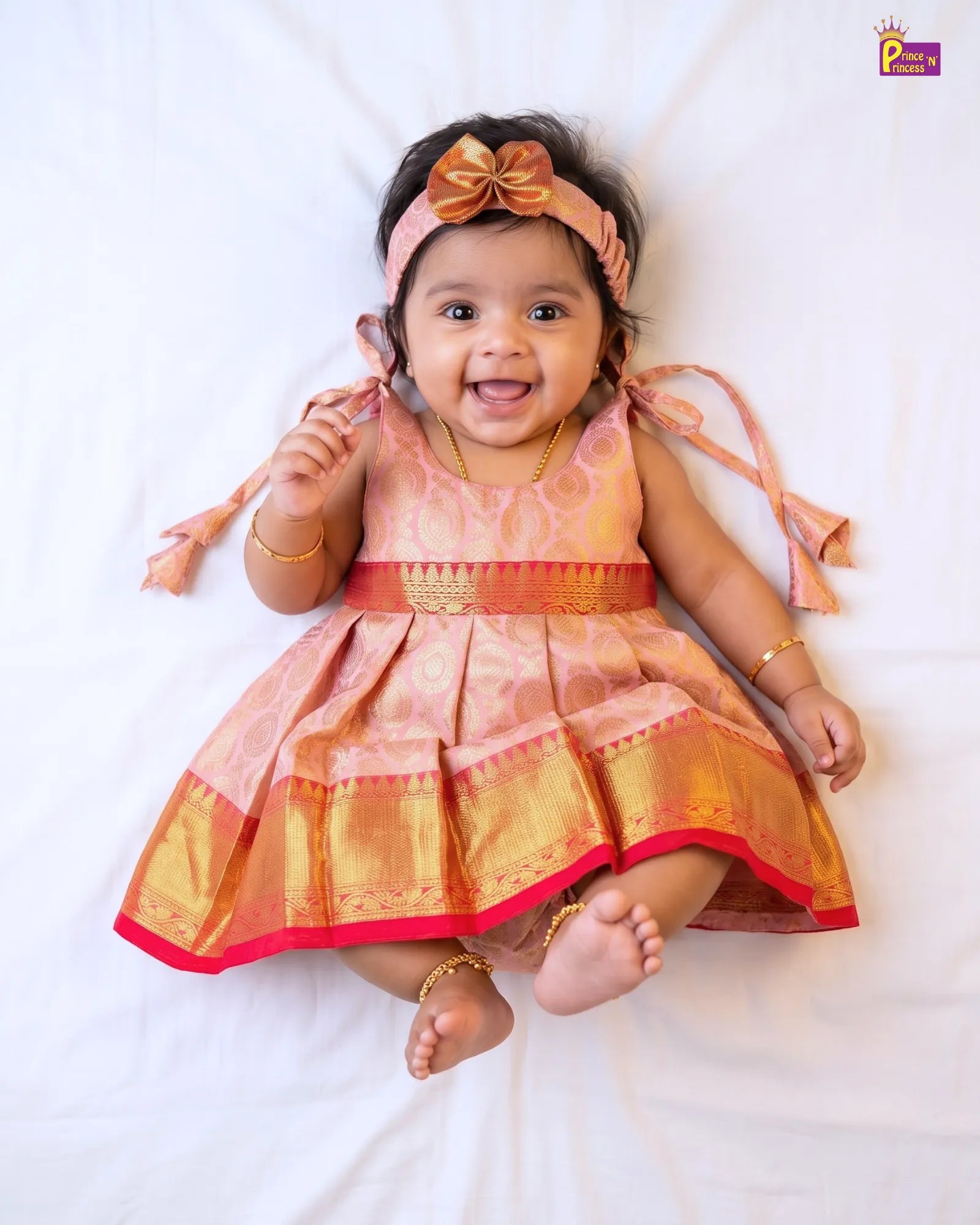 A baby girl lying on a white sheet, wearing a peach and gold traditional Indian dress with a matching bow headband, gold jewelry, and ankle bracelets.