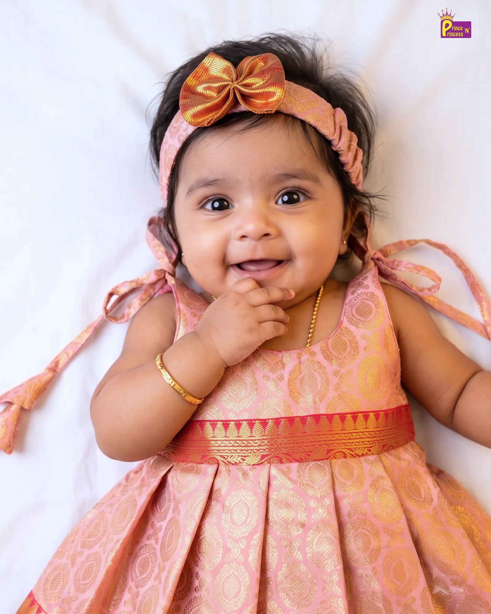 A smiling baby girl wearing a peach and gold traditional Indian dress with a matching bow headband, lying on a white fabric background.