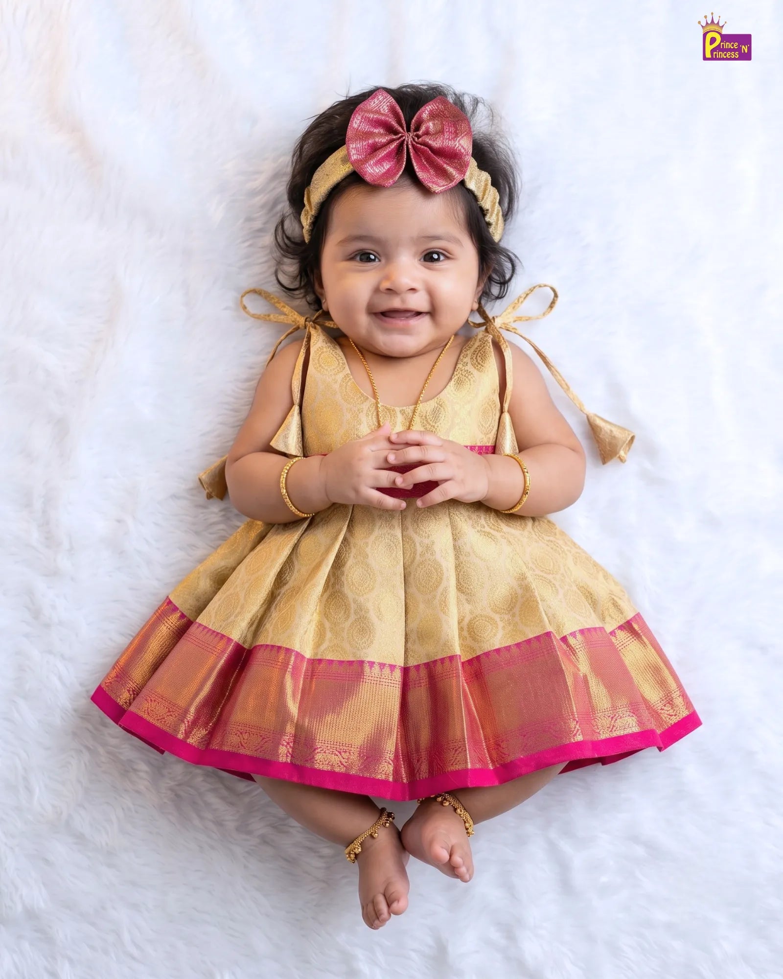 A baby girl wearing a gold and pink traditional Indian dress with a matching bow headband, sitting on a white textured blanket.