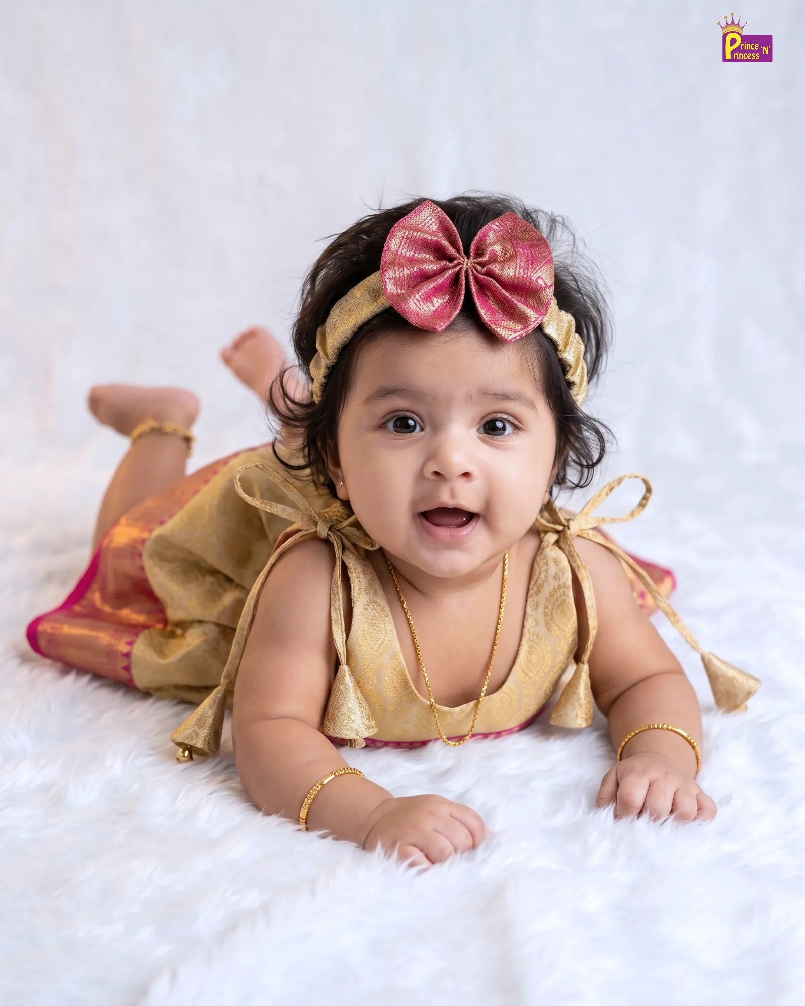 A baby girl lying on her stomach wearing a gold traditional outfit with a pink bow headband and gold jewelry, set against a white textured background.