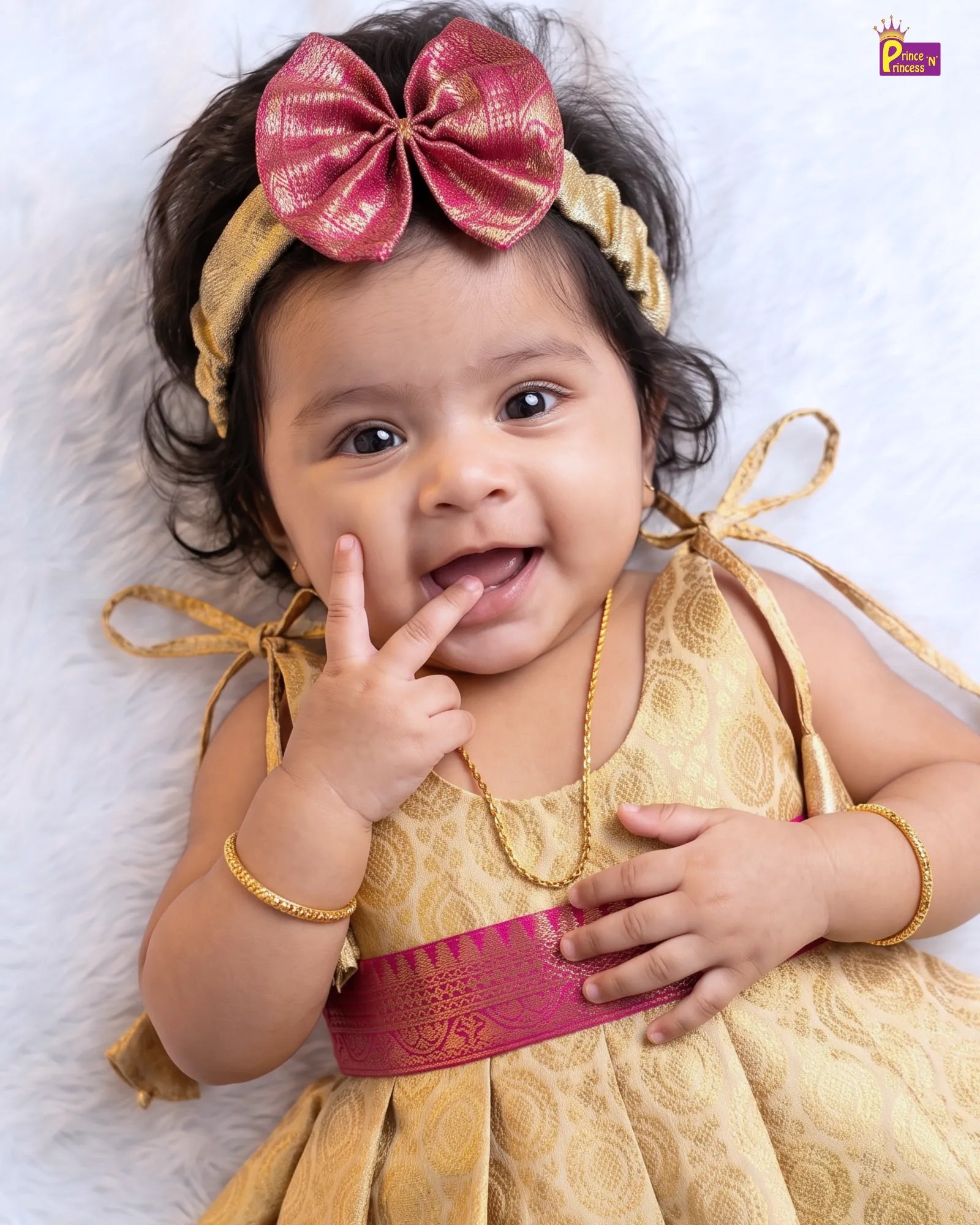 A baby girl wearing a gold patterned traditional dress with a magenta pink waist sash, a matching gold headband with a large magenta bow, and gold bangles, lying on a white faux fur surface.