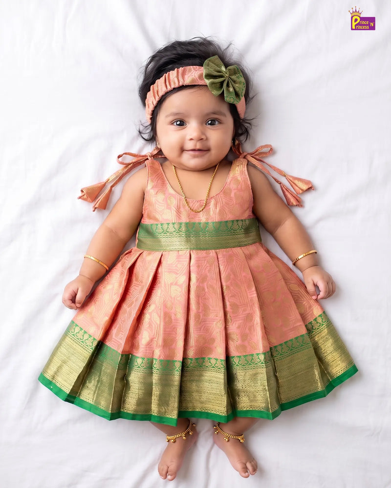 A baby girl lying on a white sheet wearing a peach and green traditional Indian dress with gold zari borders, a matching green bow headband, and gold jewelry.