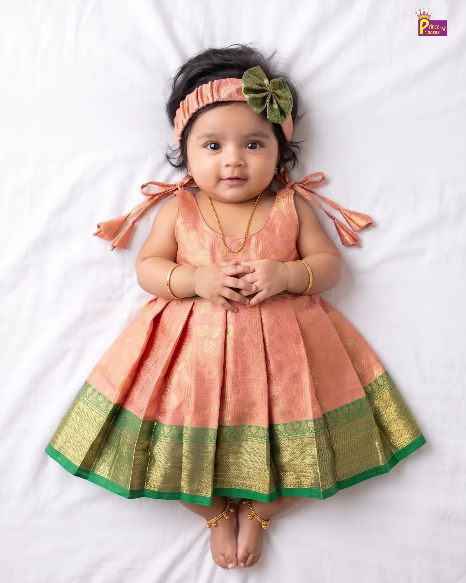 A baby girl lying on a white sheet wearing a peach and gold traditional Indian dress with a green border, a matching peach headband with a green bow, and gold jewelry.