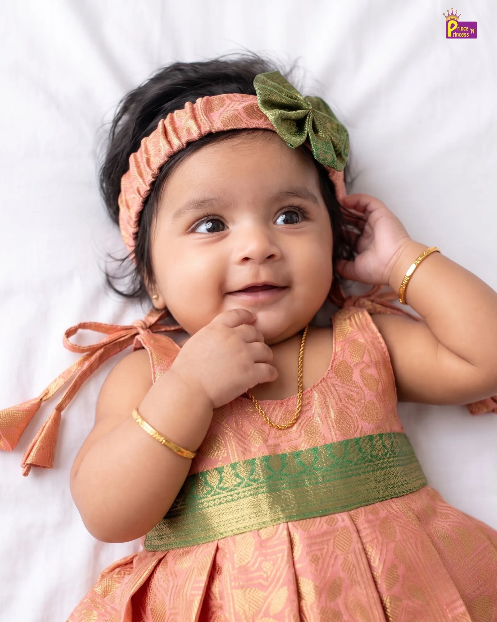 A baby girl lying on a white sheet, wearing a peach and gold patterned traditional dress with a wide green border, a green bow headband, and gold bangles.