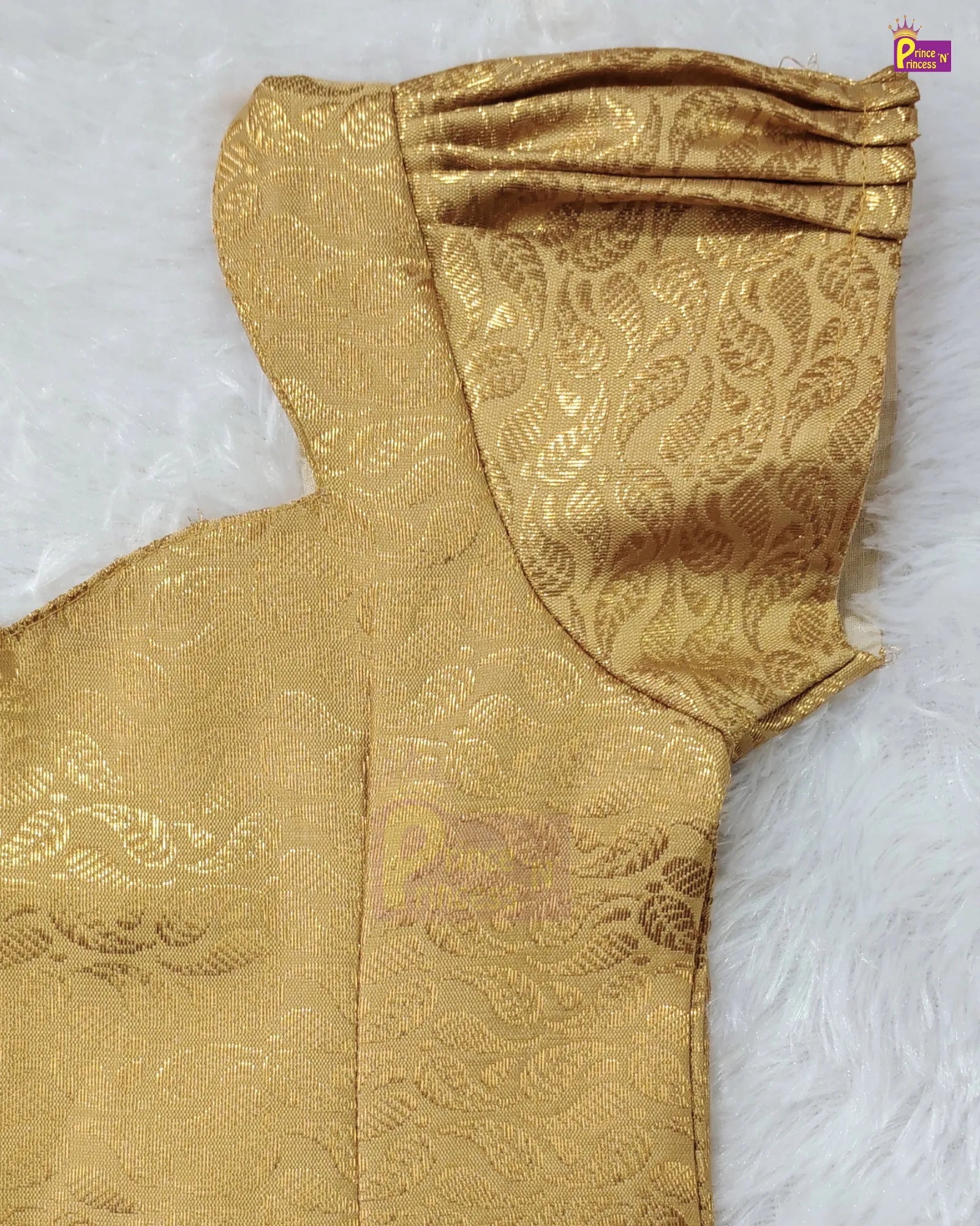 A close-up of a gold jacquard fabric garment, showing a pleated sleeve with an intricate floral pattern, laid flat on a white faux fur surface.