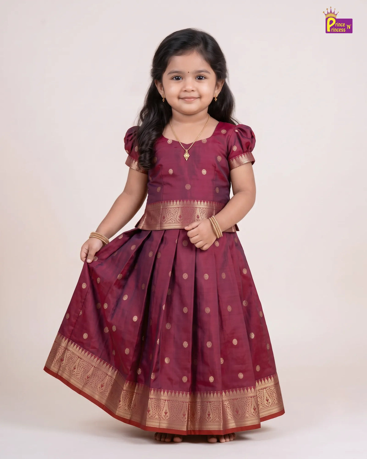 A young girl modeling a traditional Indian maroon cotton saree with gold zari work, featuring a flared skirt, short puff sleeves, and a decorative waistband, set against a plain cream background.