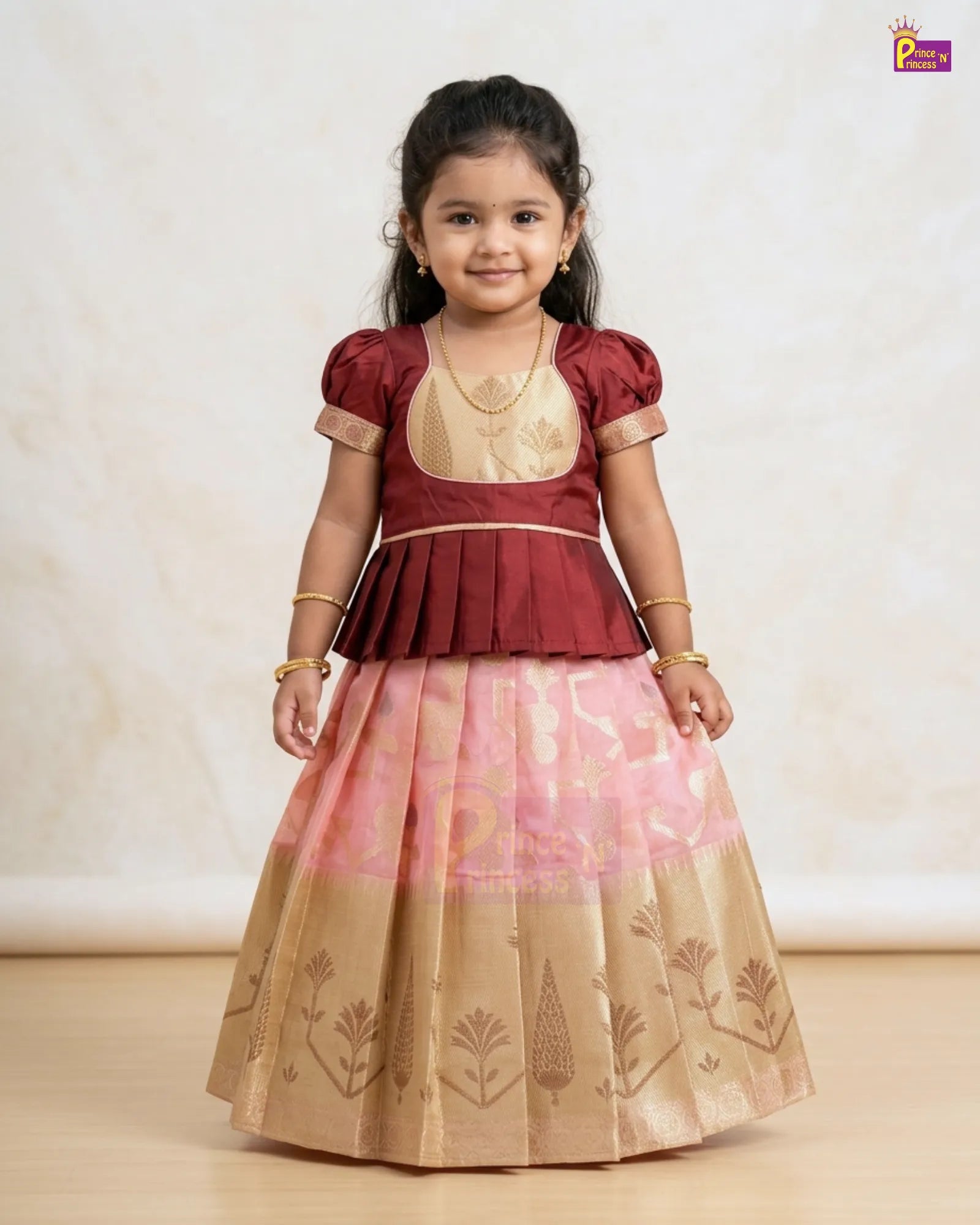 A young girl modeling a traditional Indian ethnic dress with a maroon peplum top, gold embroidered bodice, and a pink and gold patterned flared skirt, standing against a neutral beige background.
