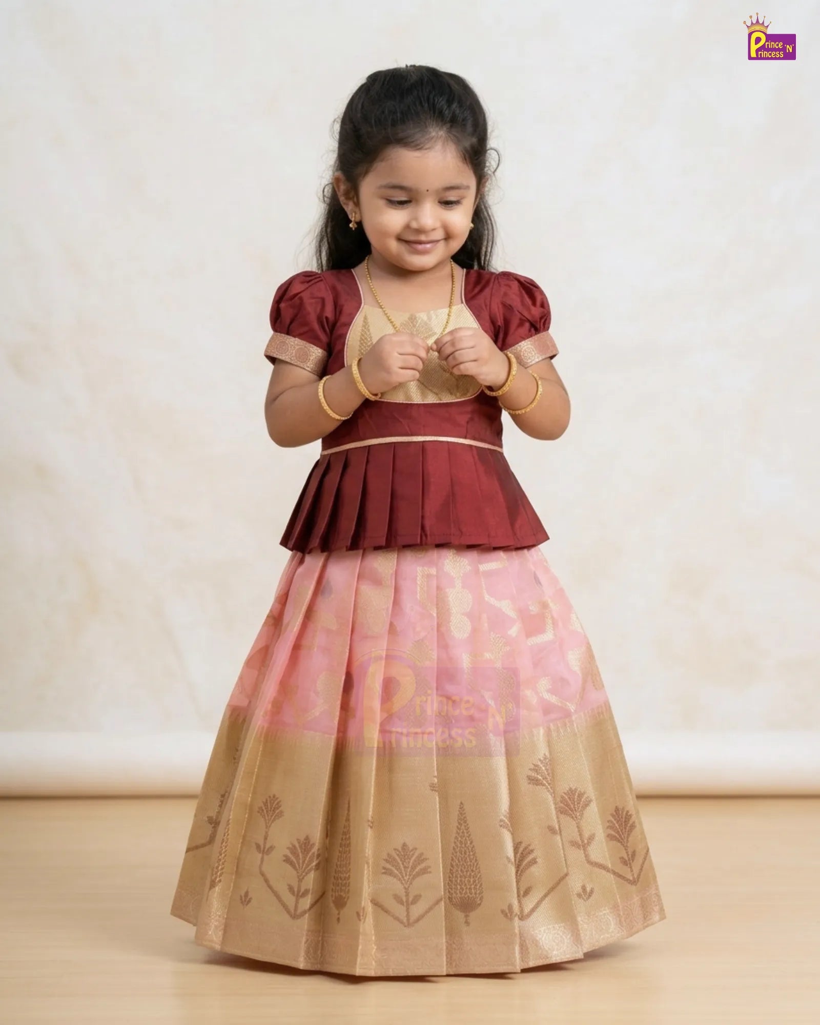 A young girl modeling a traditional Indian ethnic dress with a maroon peplum-style bodice, a light pink upper skirt, and a beige lower skirt with printed motifs, set against a neutral cream background.
