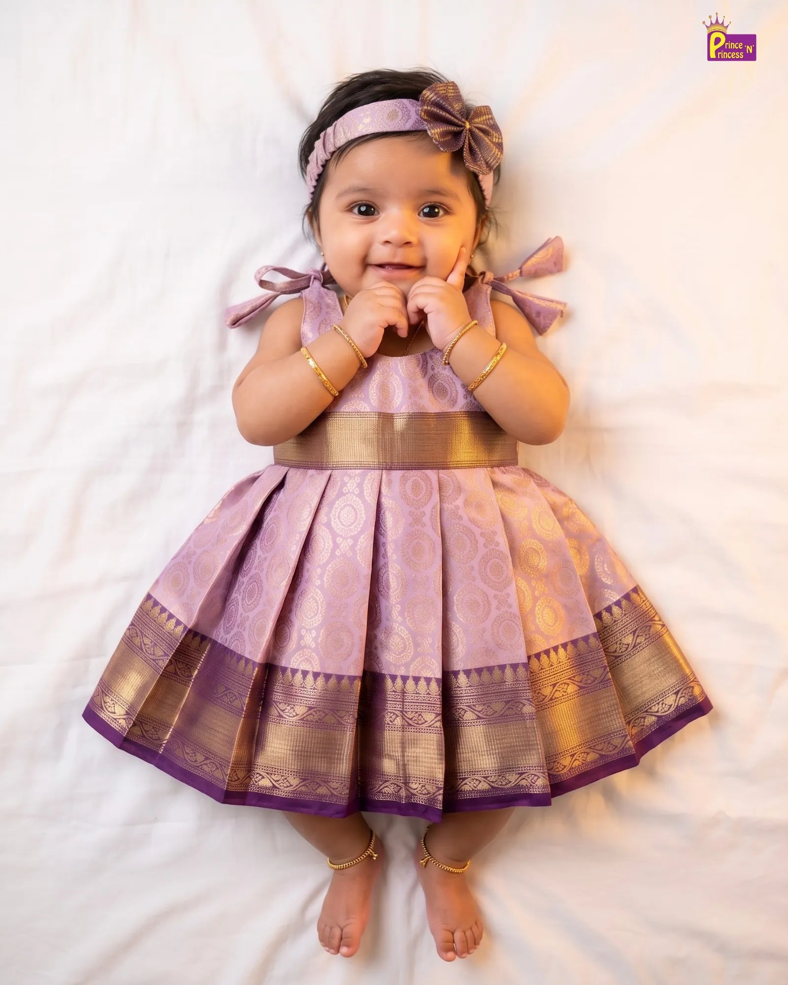 A baby girl lying on a white sheet, wearing a lavender and gold traditional Indian dress with a pleated skirt, a matching headband with a bow, and gold bangles and ankle bracelets.