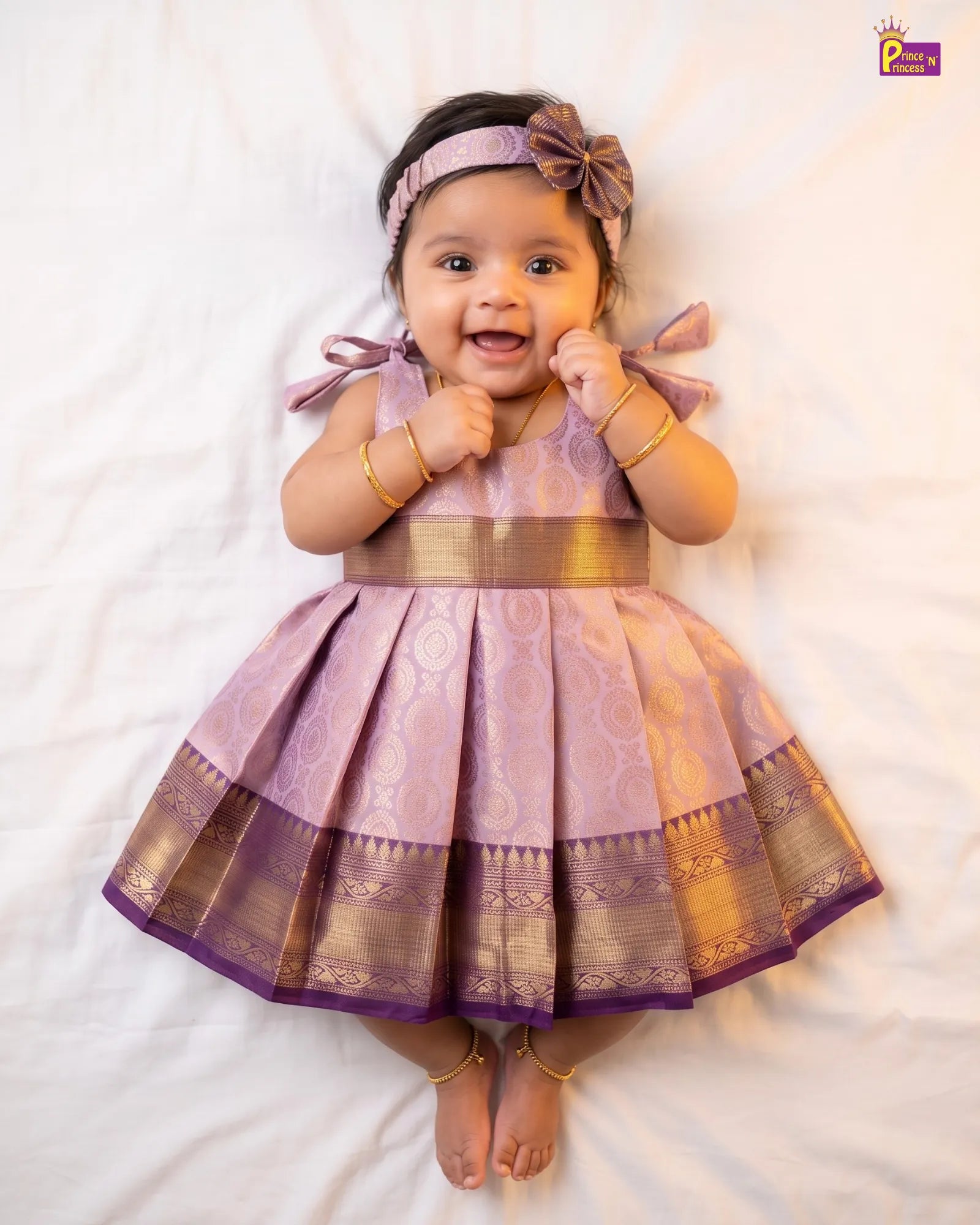 A baby girl lying on a white sheet wearing a lavender and gold traditional Indian dress with a matching headband and bow.