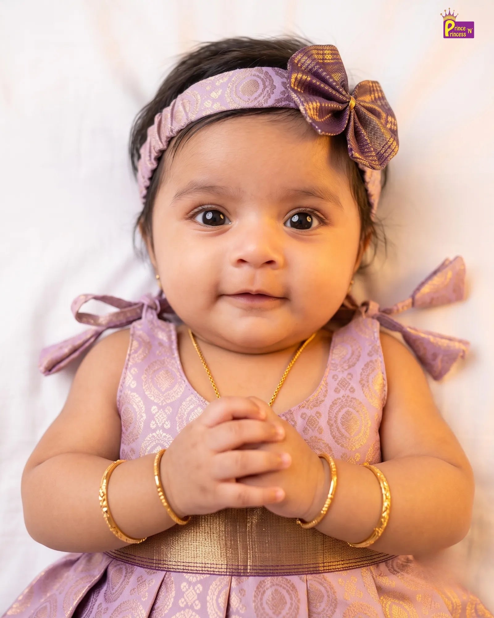 A baby girl wearing a lavender and gold patterned dress, matching headband with a bow, and gold bangles, lying on a white background.
