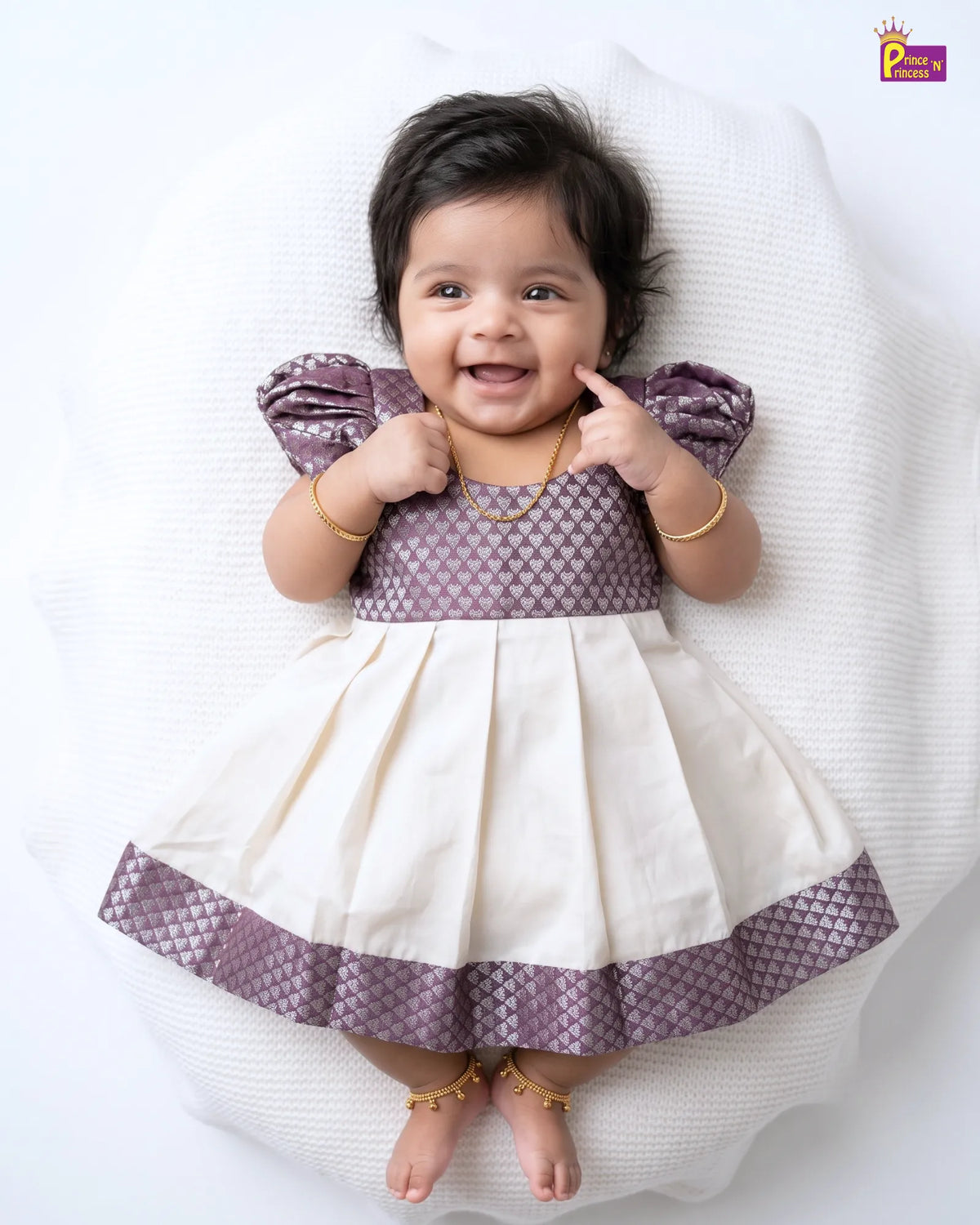 A baby girl lying on a white textured blanket, wearing a purple and cream dress with gold jewelry.