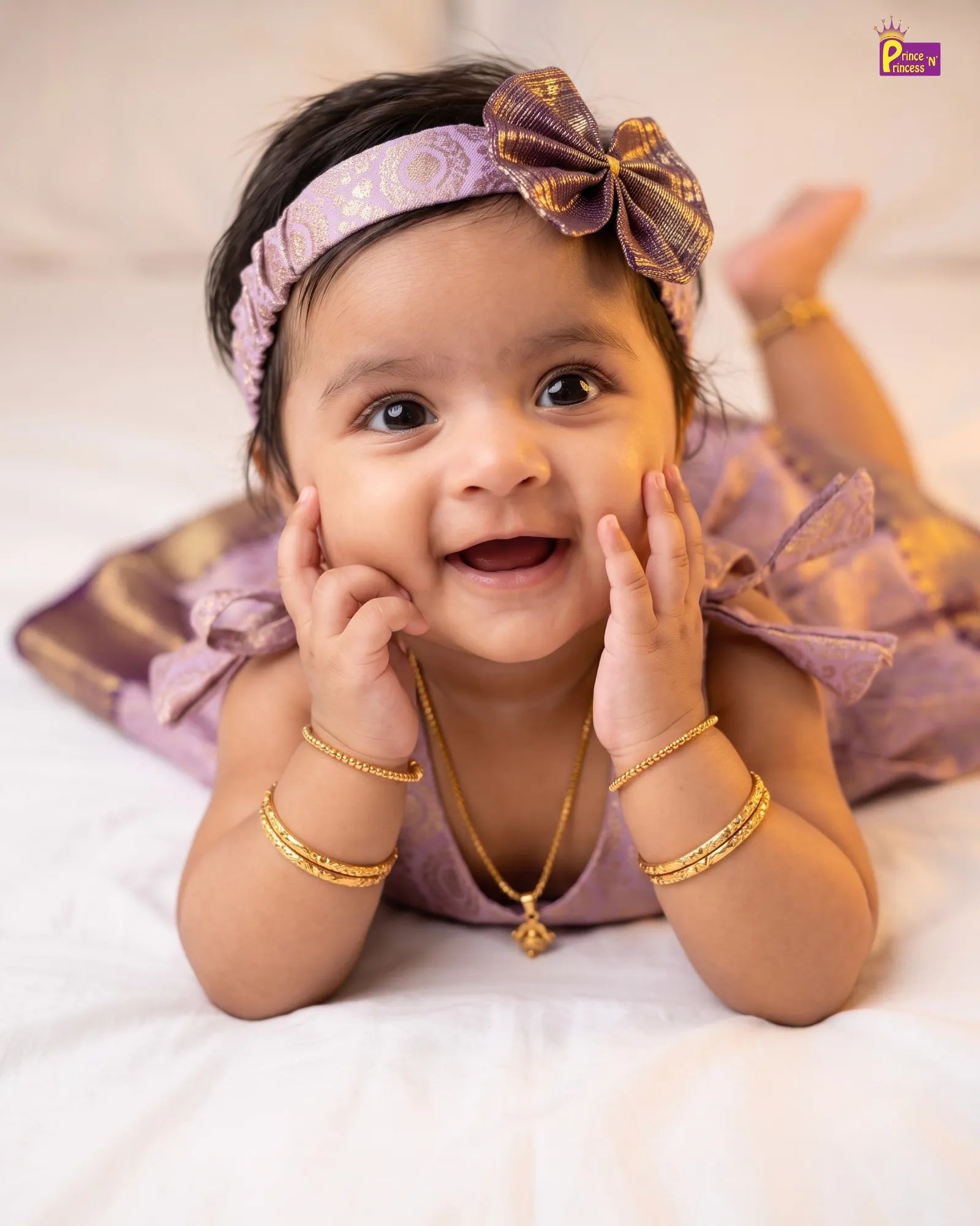 A baby lying on her stomach wearing a lavender and gold traditional outfit with a matching bow headband, gold bangles, and a gold necklace, posing on a white bed.