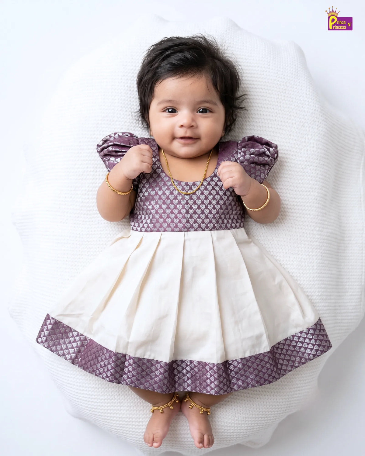 A baby girl lying on a white textured cushion, wearing a purple and cream tiered dress with gold jewelry.