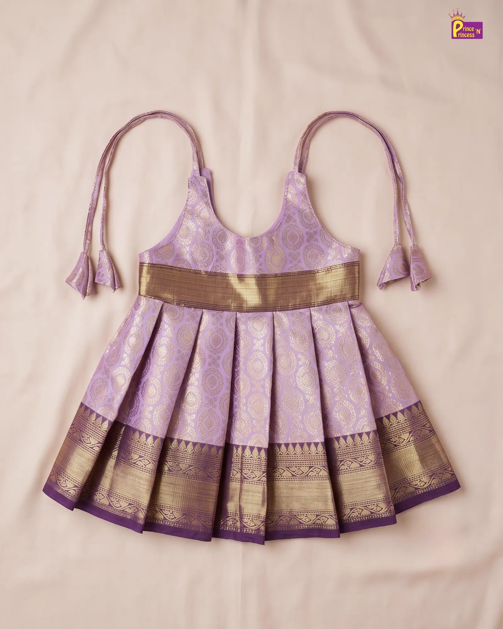 A purple and gold traditional Indian toddler dress with a pleated skirt, halter straps, and a wide gold waistband, laid flat on a light beige fabric background.