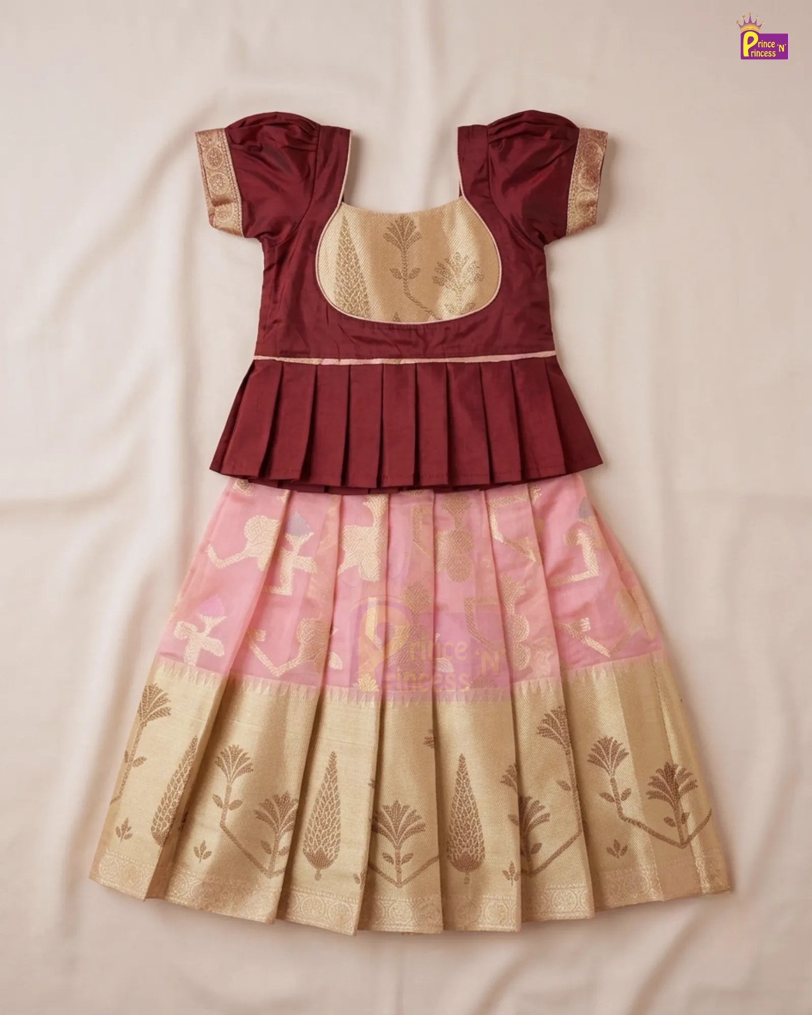 A traditional Indian girl's dress with a maroon peplum top, beige embroidered bodice, and a pleated skirt in pink and beige with gold zari motifs, laid flat on a light beige fabric background.